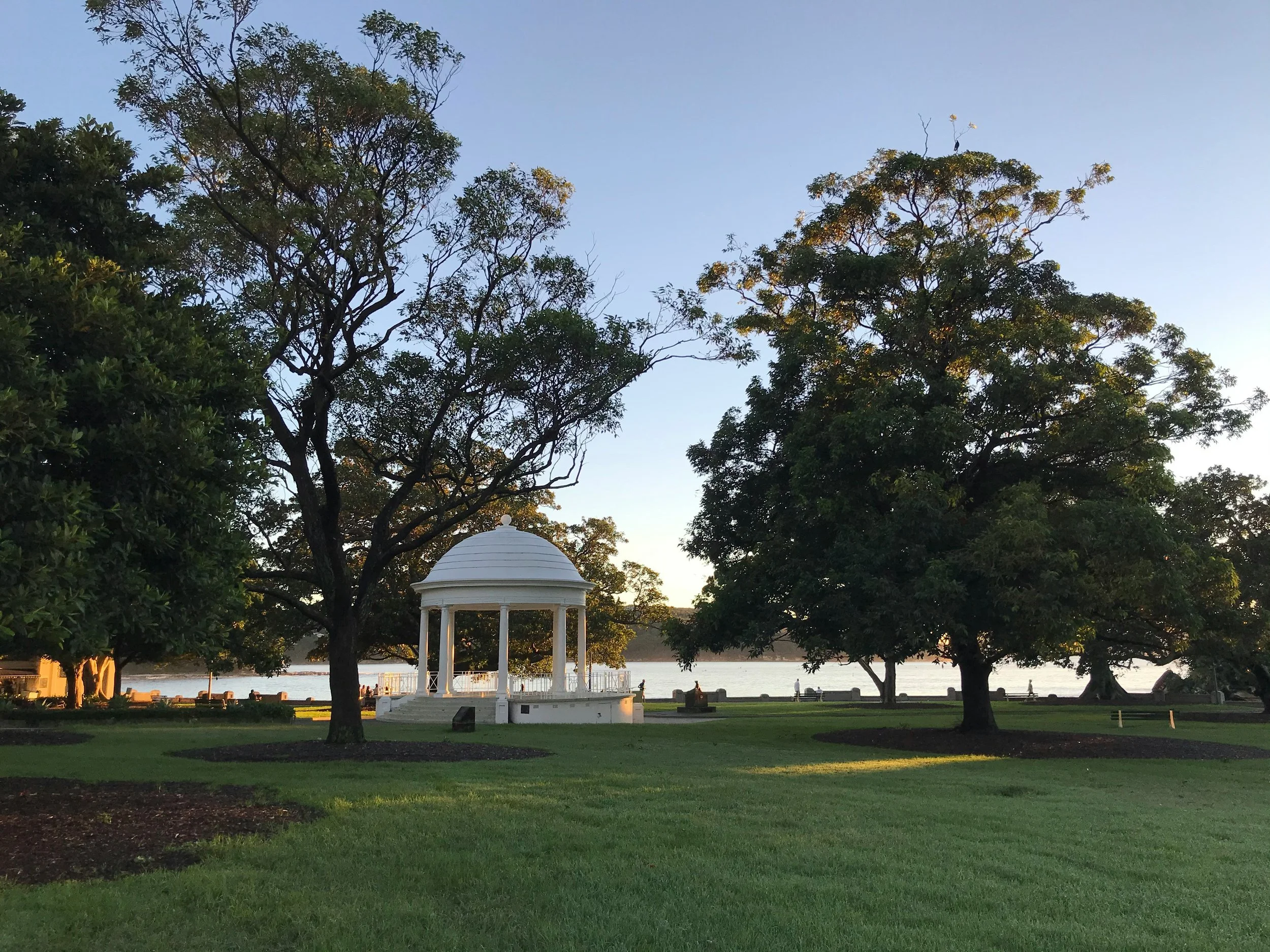 A park with green grass, large trees, and a white gazebo near a body of water during sunset.
