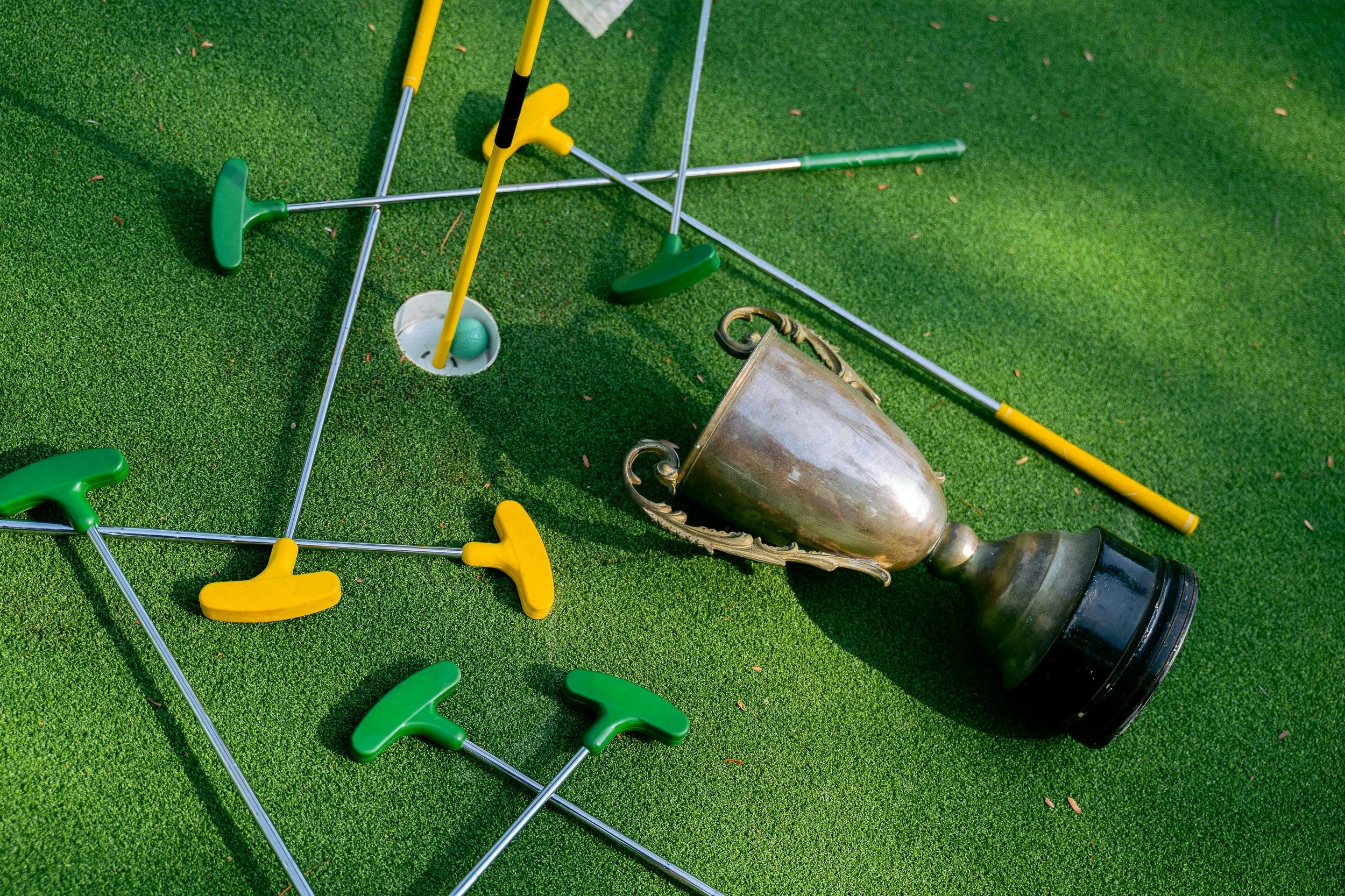 A golf trophy lying on the green, surrounded by scattered golf clubs with green and yellow handles on artificial grass.