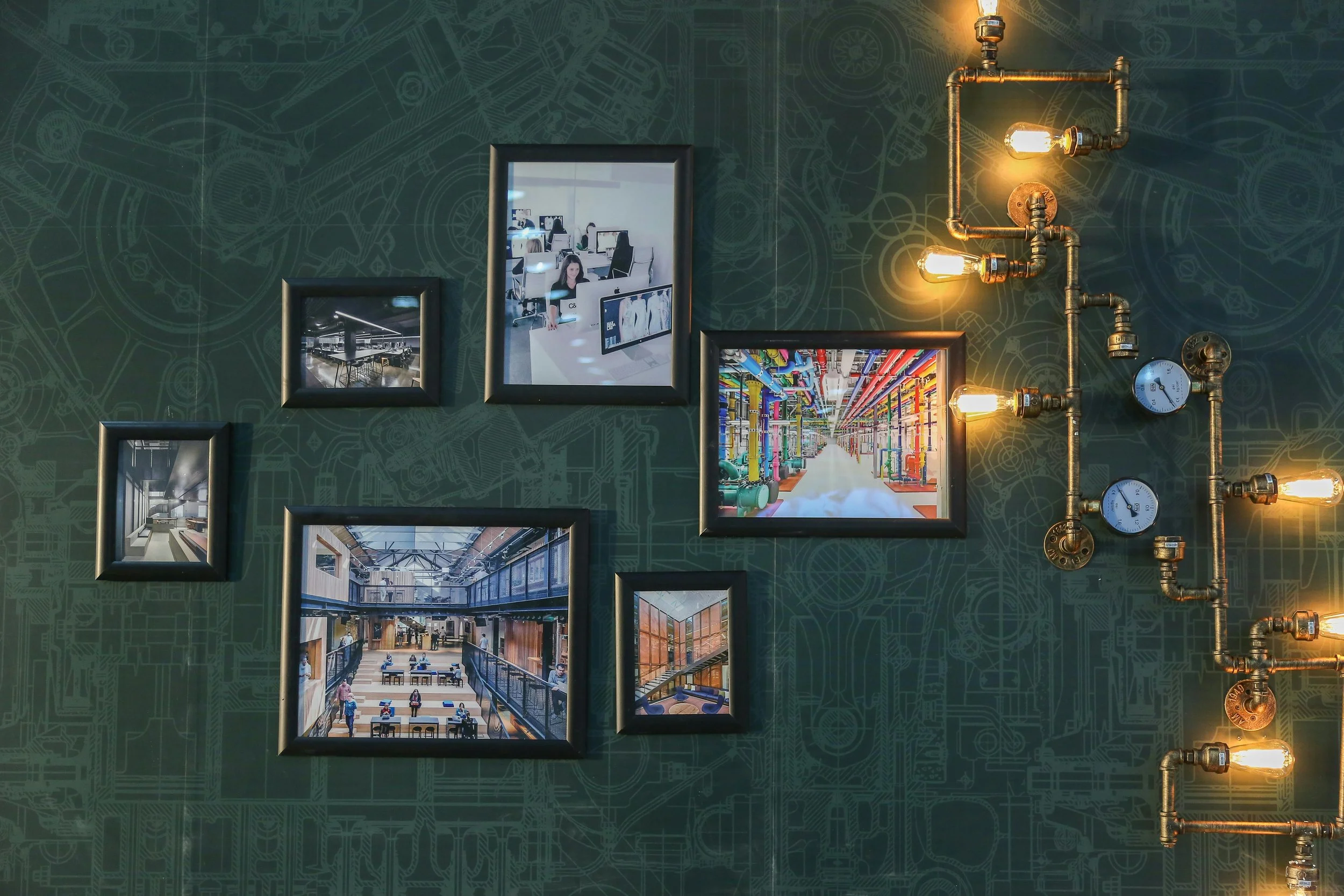 Gallery wall with framed photos of modern workspaces and industrial interiors, next to a pipe system with exposed bulbs and gauges on a dark patterned wall.