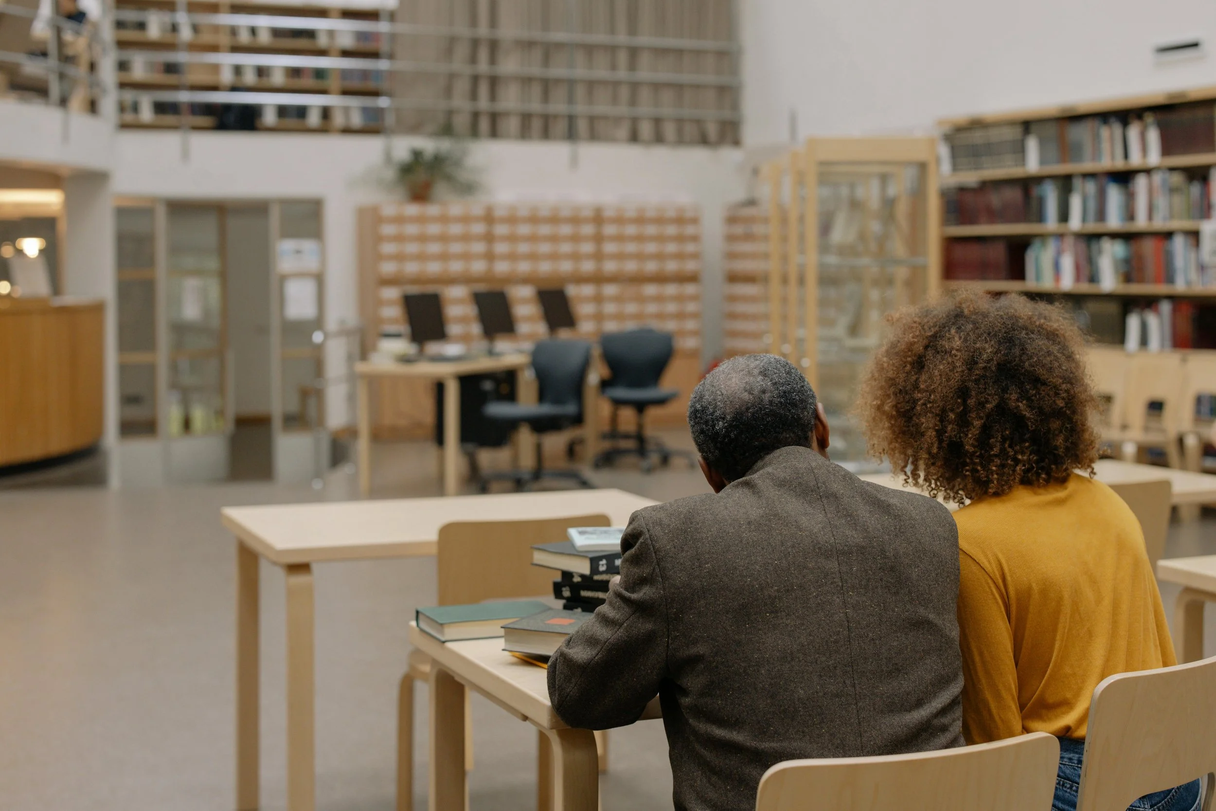 Two people sit with their backs to the camera at a table in a library, facing empty chairs and bookshelves.
