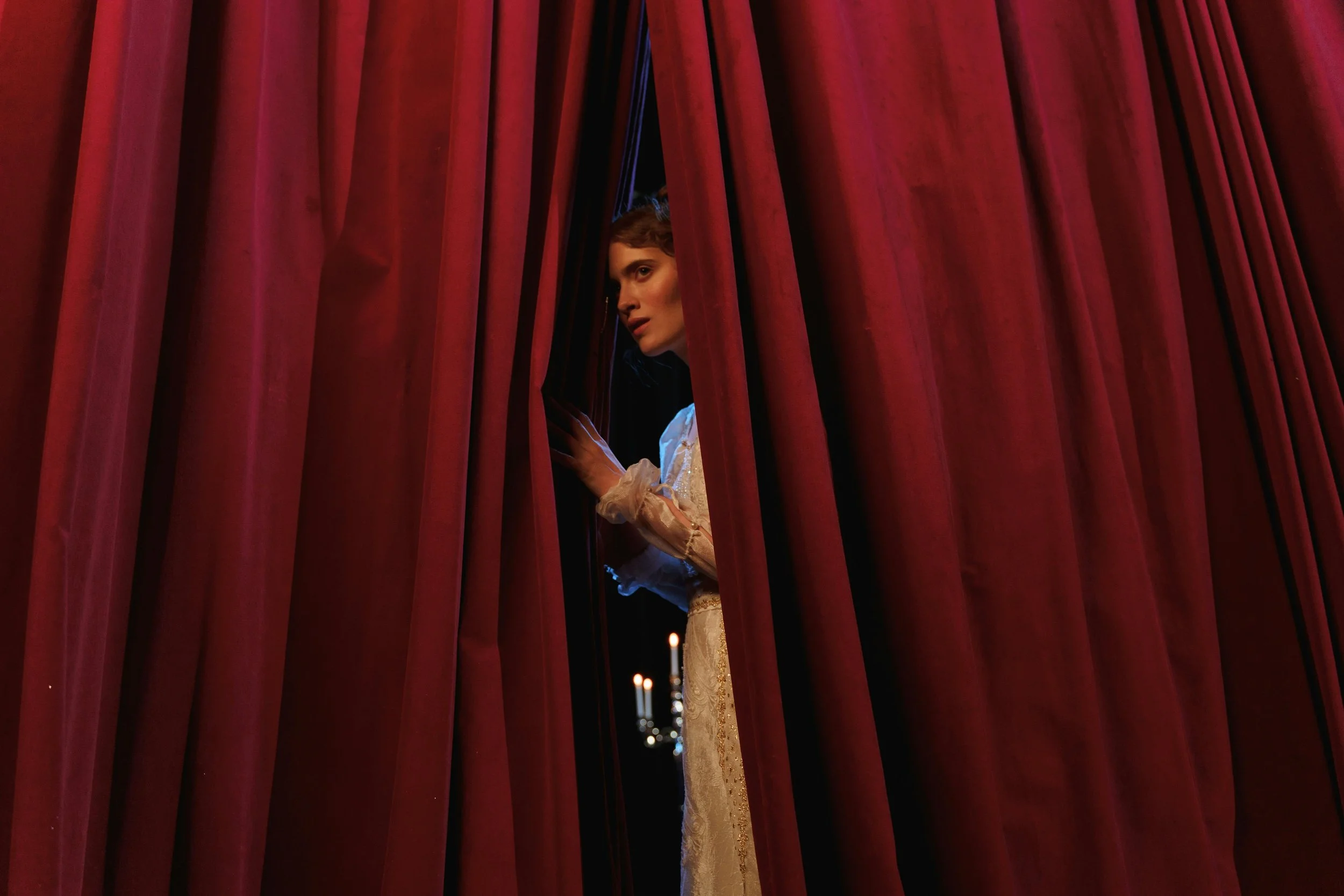 A woman peering through red stage curtains, wearing a vintage lace dress, with a candelabra in the background.