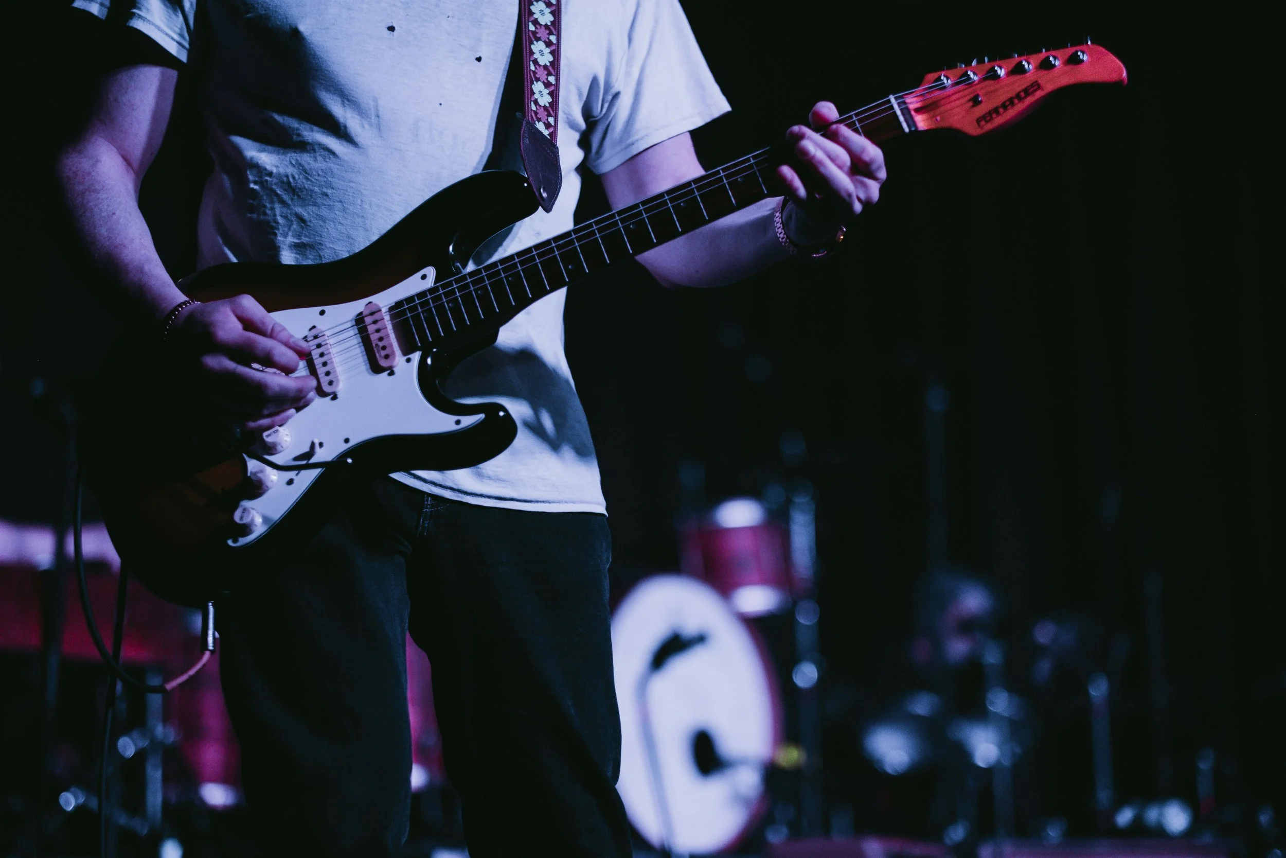 Person playing an electric guitar on stage with a drum set in the background, dark lighting with focus on the guitar.
