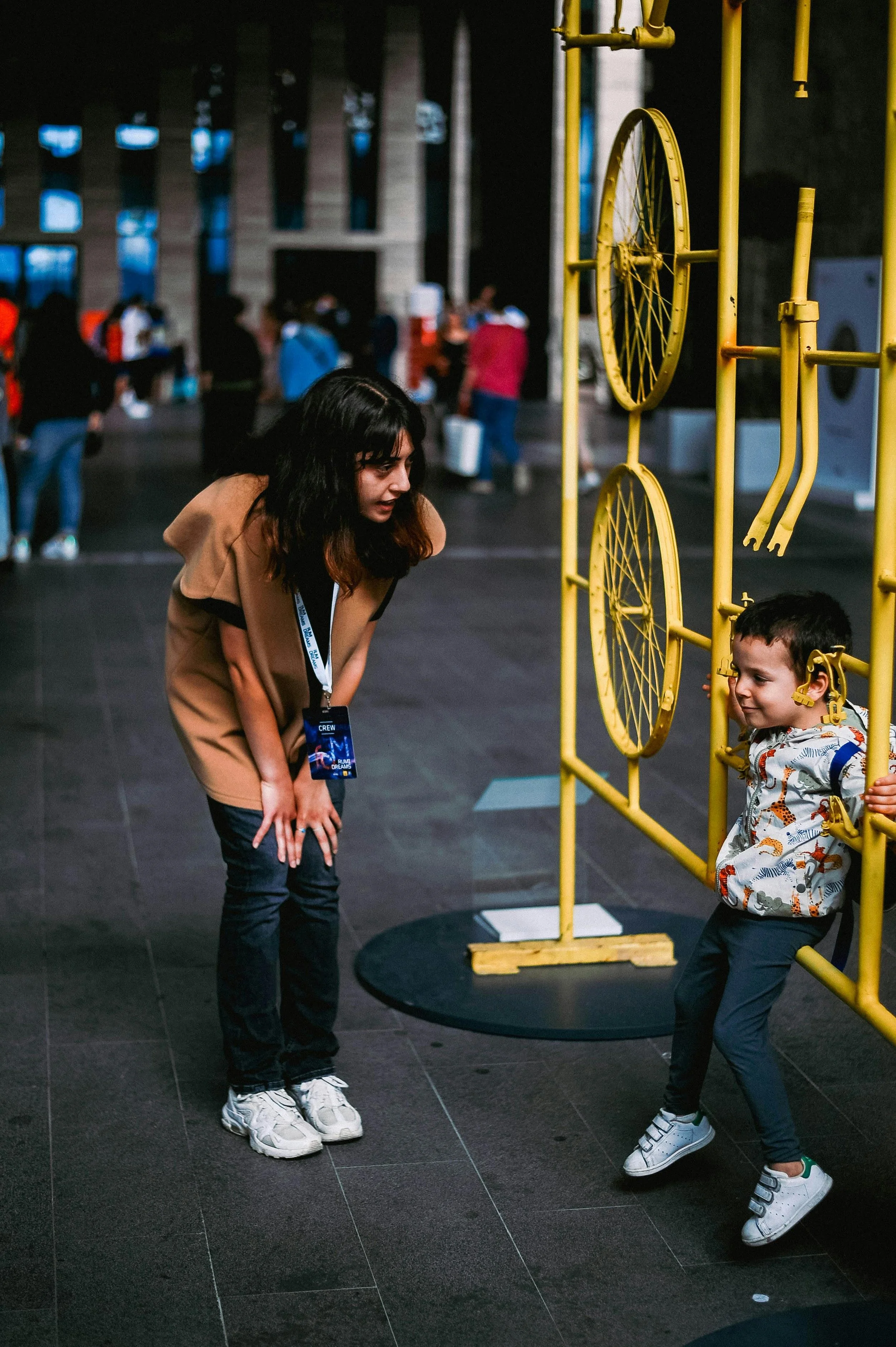 A woman bending forward is talking to a young boy who is smiling and holding onto a yellow climbing frame. The background shows an indoor space with several people walking and standing.