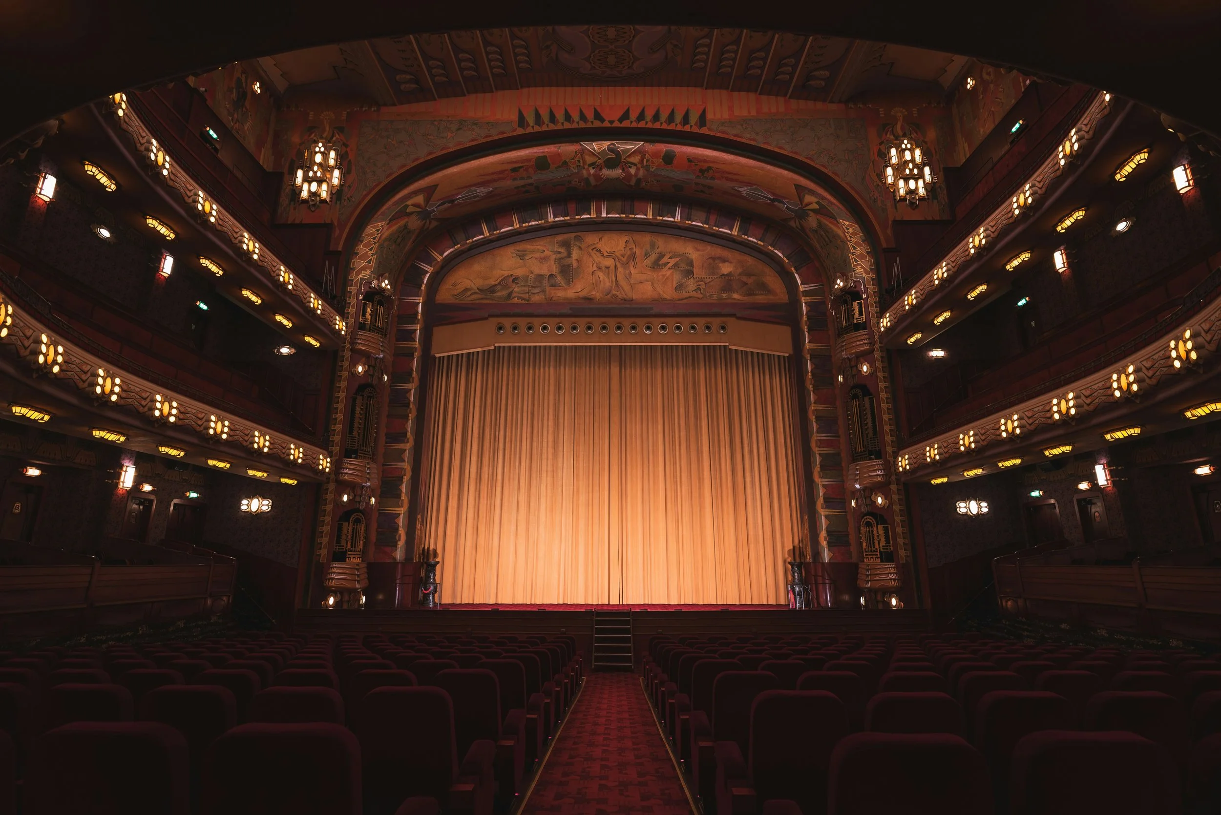 Empty theater auditorium with closed golden curtains, ornate interior decor, multiple balconies with lights, and empty seats facing the stage.