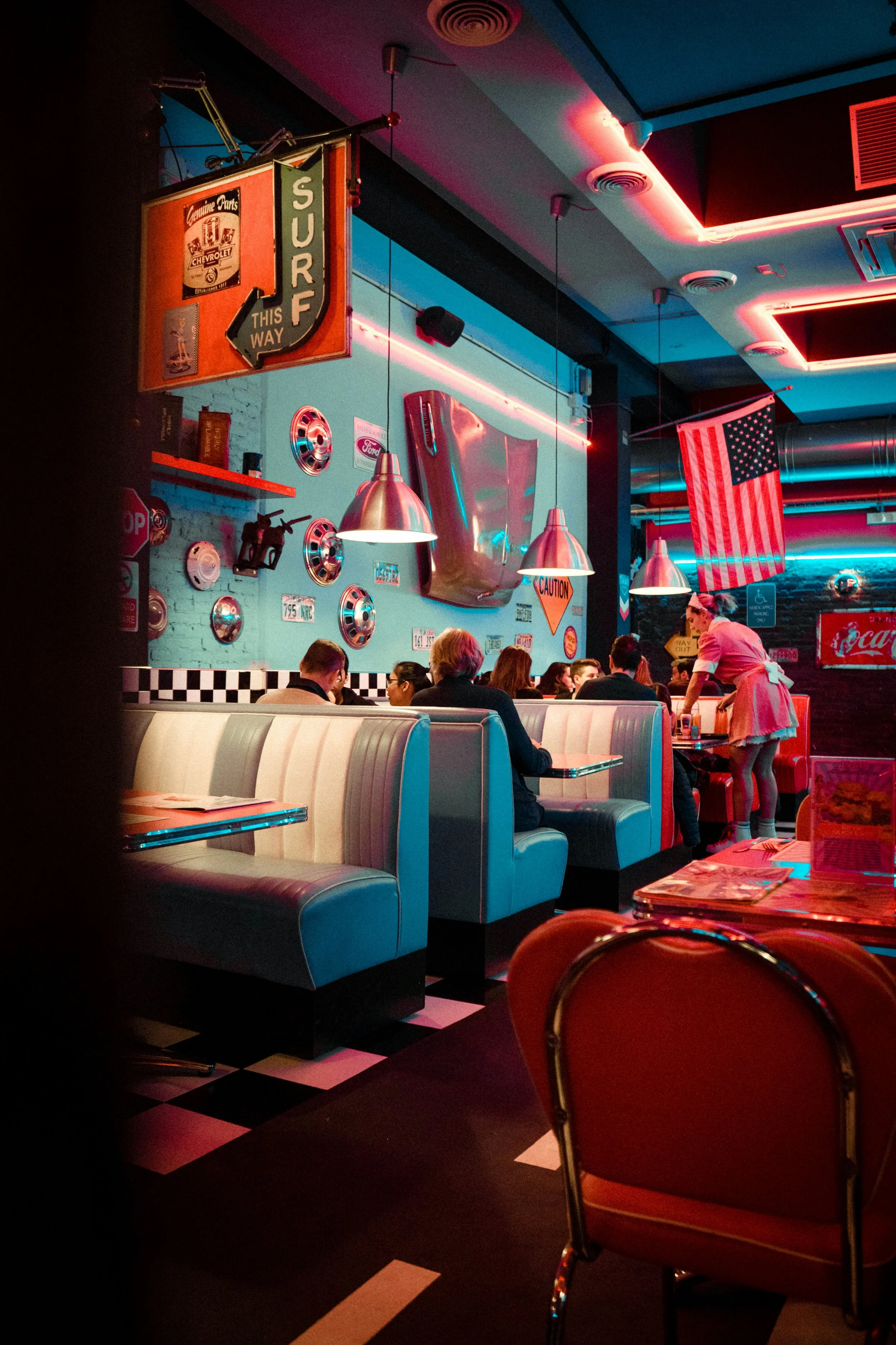 Diners sitting in a retro-style American diner with vintage car parts and neon lights decor, waitresses serving food under hanging pendant lights, American flag in the background, black-and-white checkered floor, and 1950s themed memorabilia on the walls.