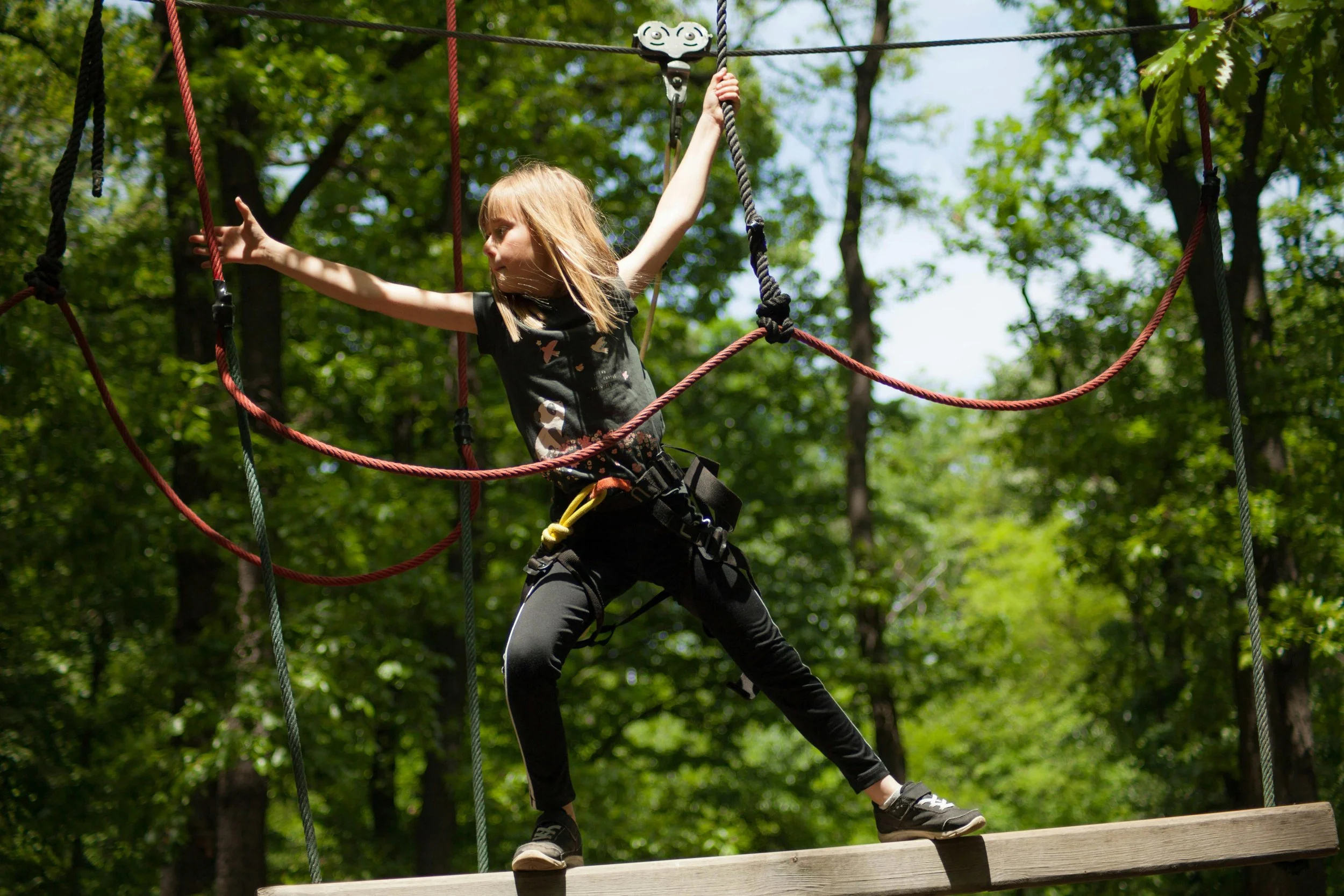 A young girl wearing a harness walking on a wooden platform in a treetop adventure course surrounded by green trees.