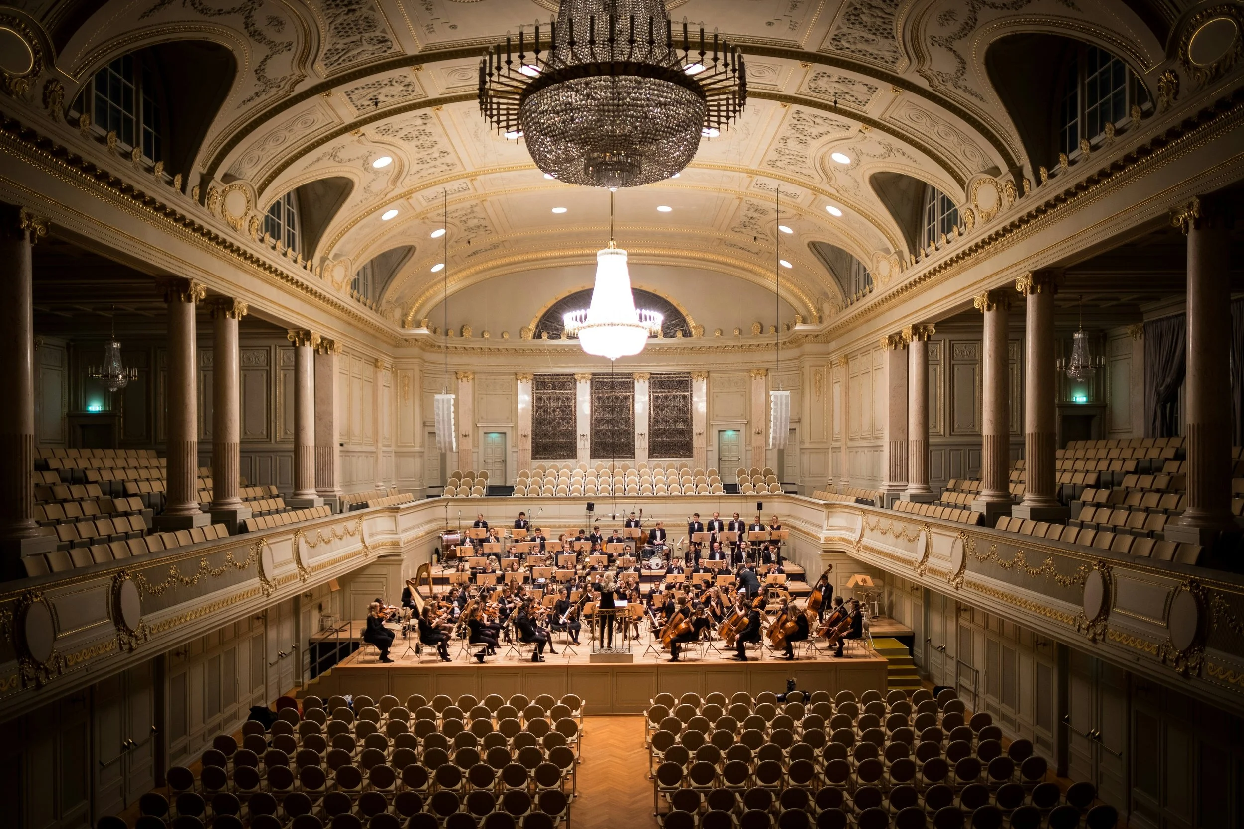 Orchestra performing in a grand, ornate hall with high vaulted ceiling, chandeliers, and rows of empty seats. Curated Event Listings in Columbia, Maryland