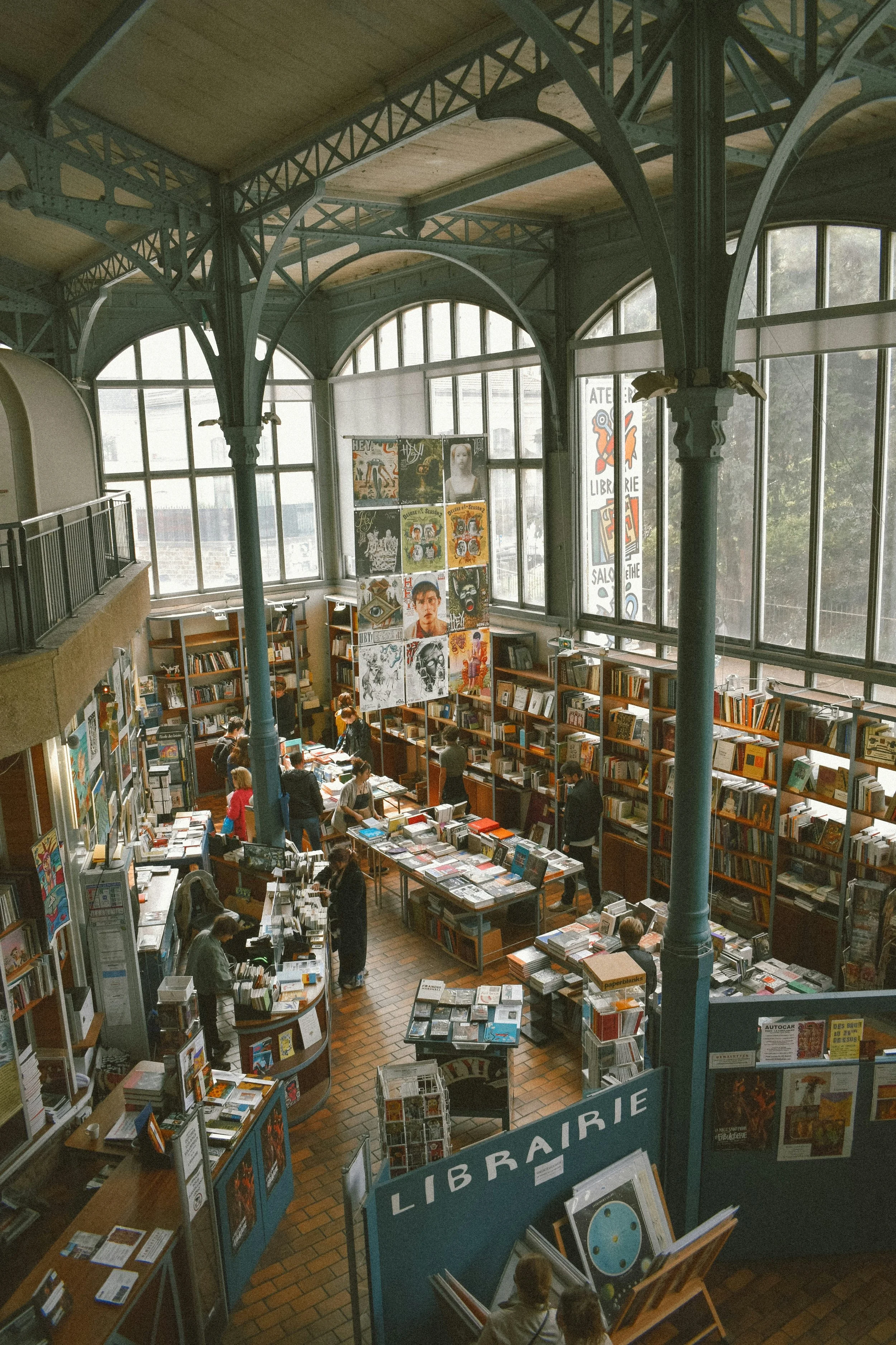 Inside a large, vintage-style bookstore with tall windows, metal framework, and bookshelves filled with books and artwork. Customers browse and shop.