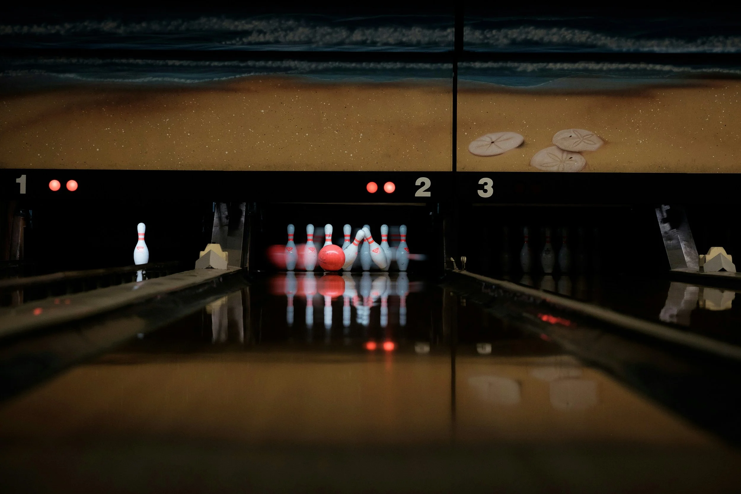 Bowling alley with pins set up at the end of the lane, pins are white with red stripes, and a red bowling ball is approaching them. The background shows a mural of a beach with sand and shells.