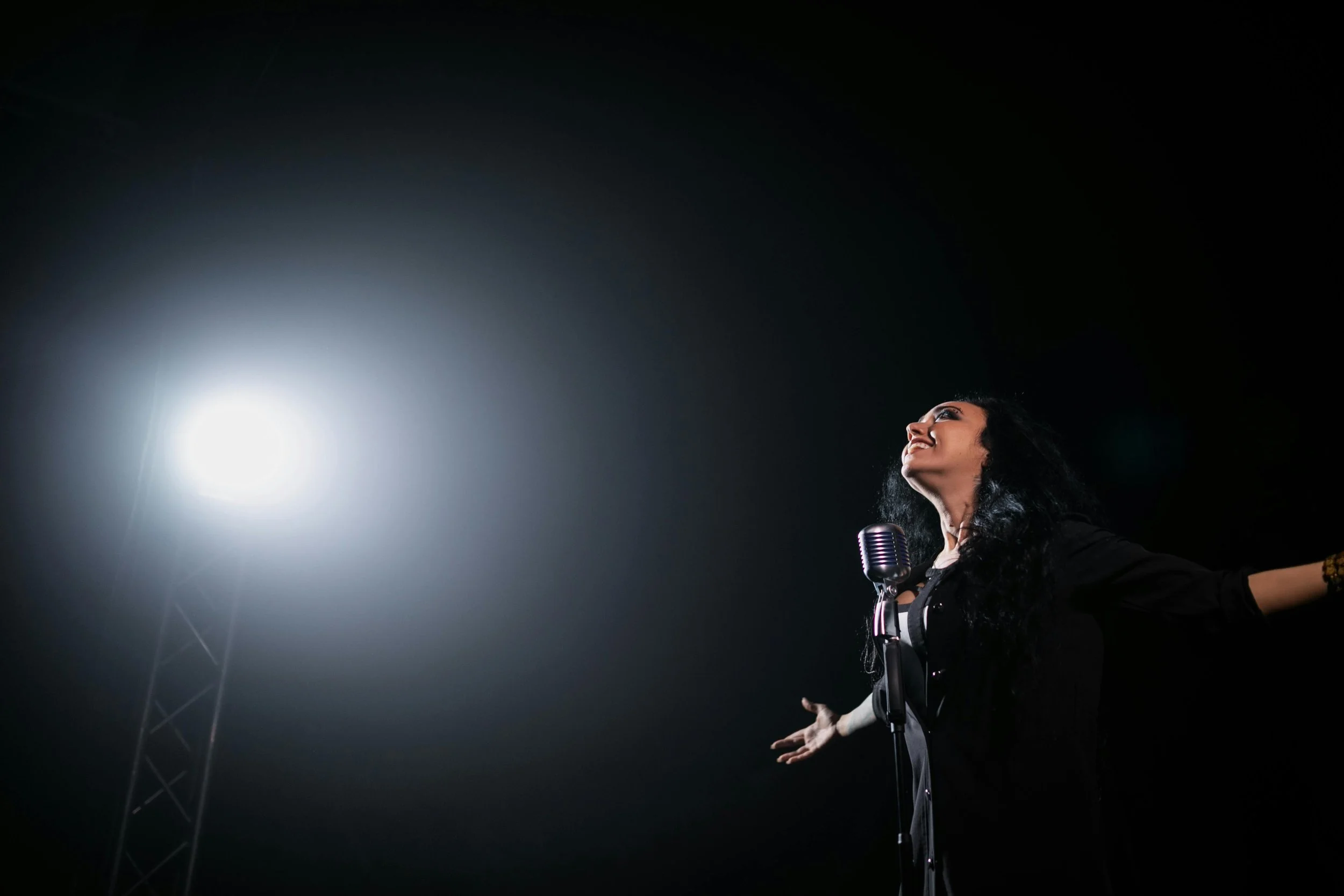 A woman singing on stage with a vintage microphone, arms outstretched, illuminated by a bright spotlight against a dark background. Curated Event Listings in Columbia, Maryland