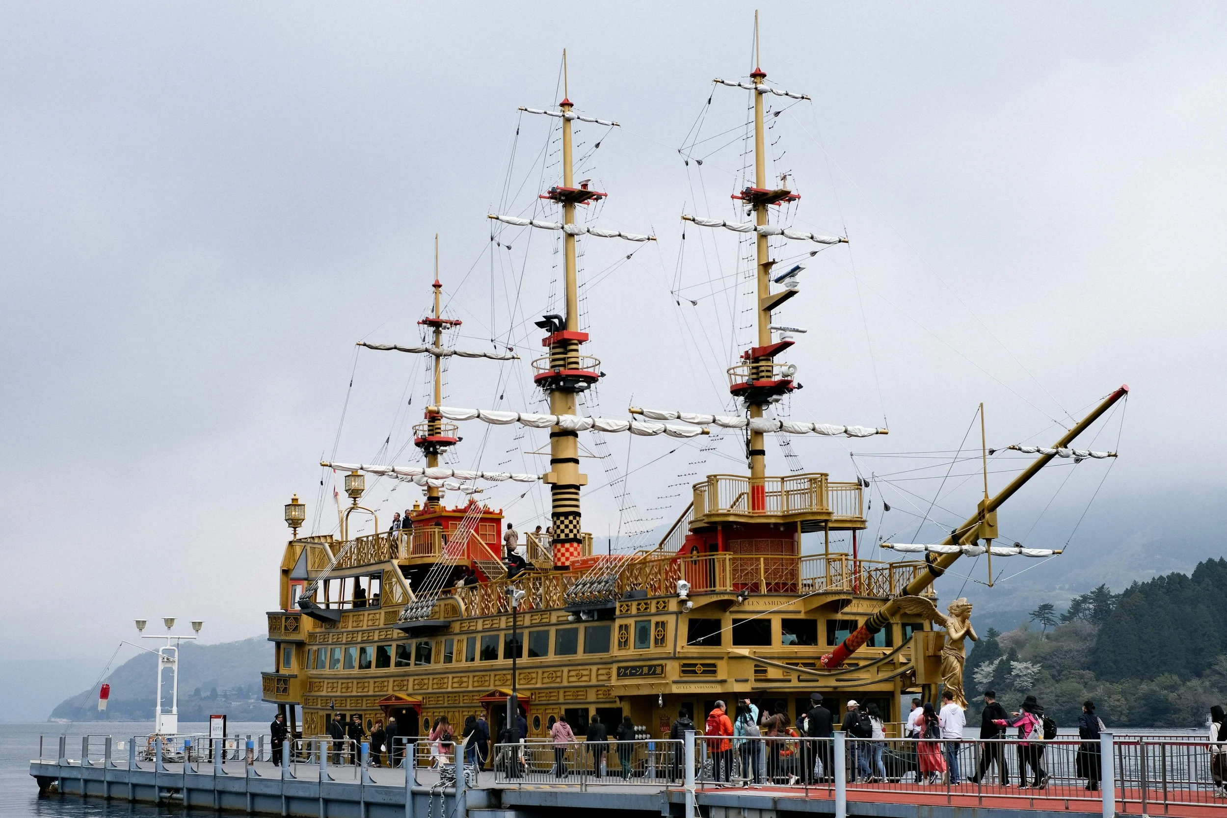 The image shows a large, colorful pirate ship-themed attraction docked at a pier with people walking along the pier. The ship has multiple masts with sails and rigging, and it is painted in gold, red, and black colors with decorative details. The background features overcast sky, mountains, and water.