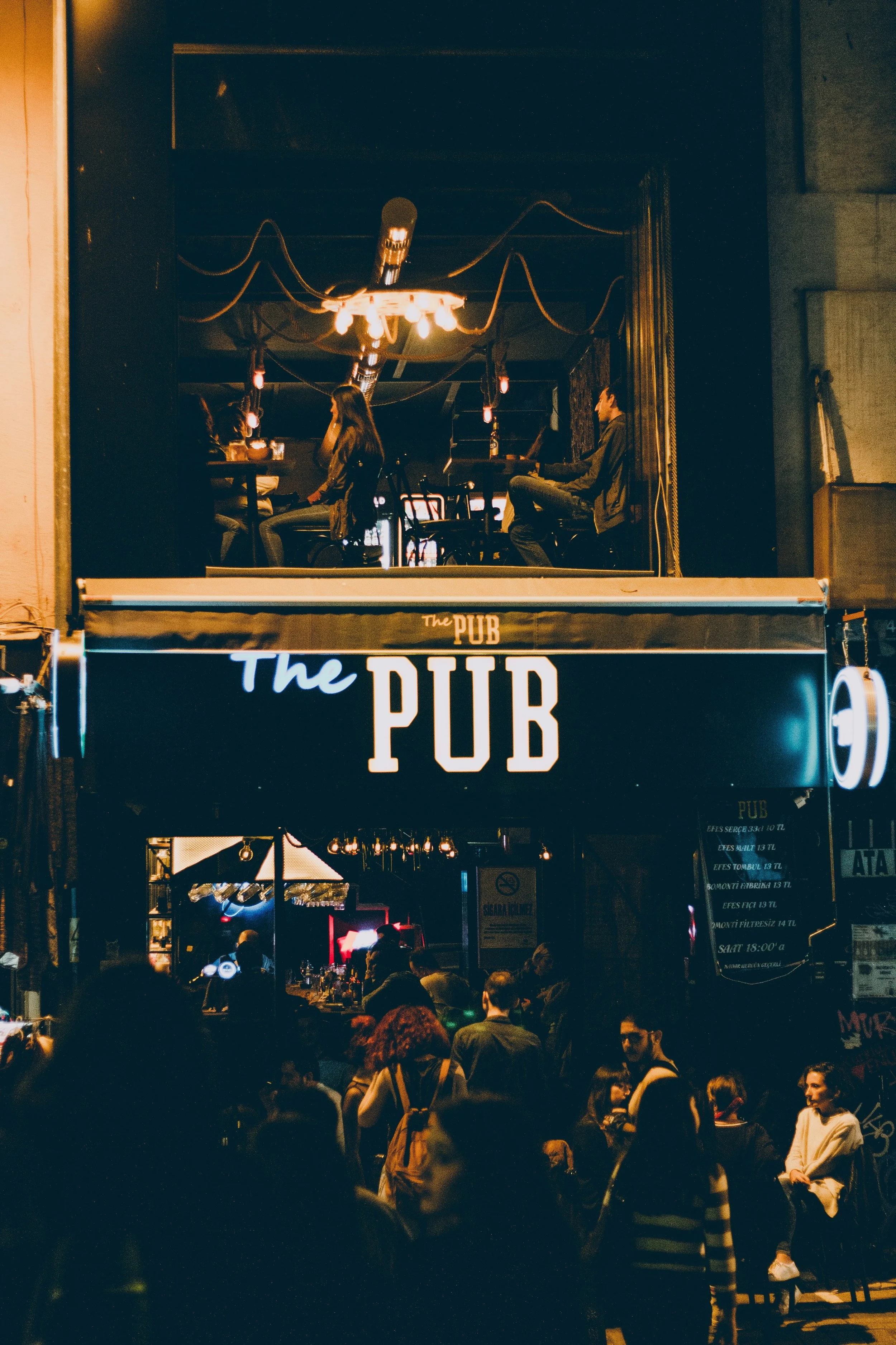 Nighttime scene outside a lively pub with a crowd of people gathered near the entrance, a bar visible inside, and a two-story building with a sign reading "The PUB" above the entrance.