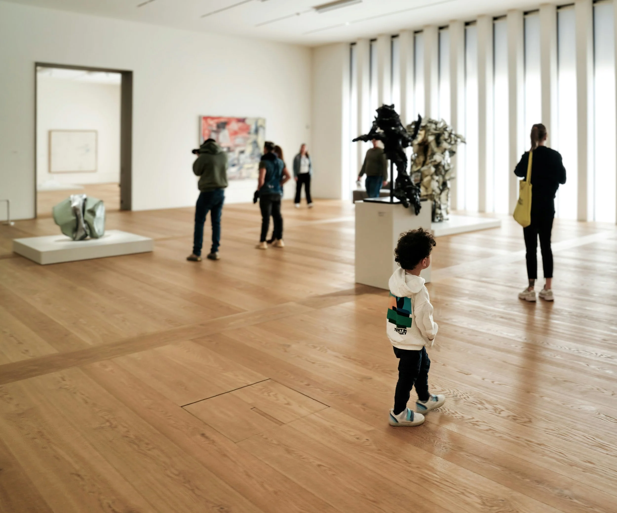 A young boy in a white hoodie walking through a spacious art gallery with wooden floors and white walls, observing sculptures and paintings.