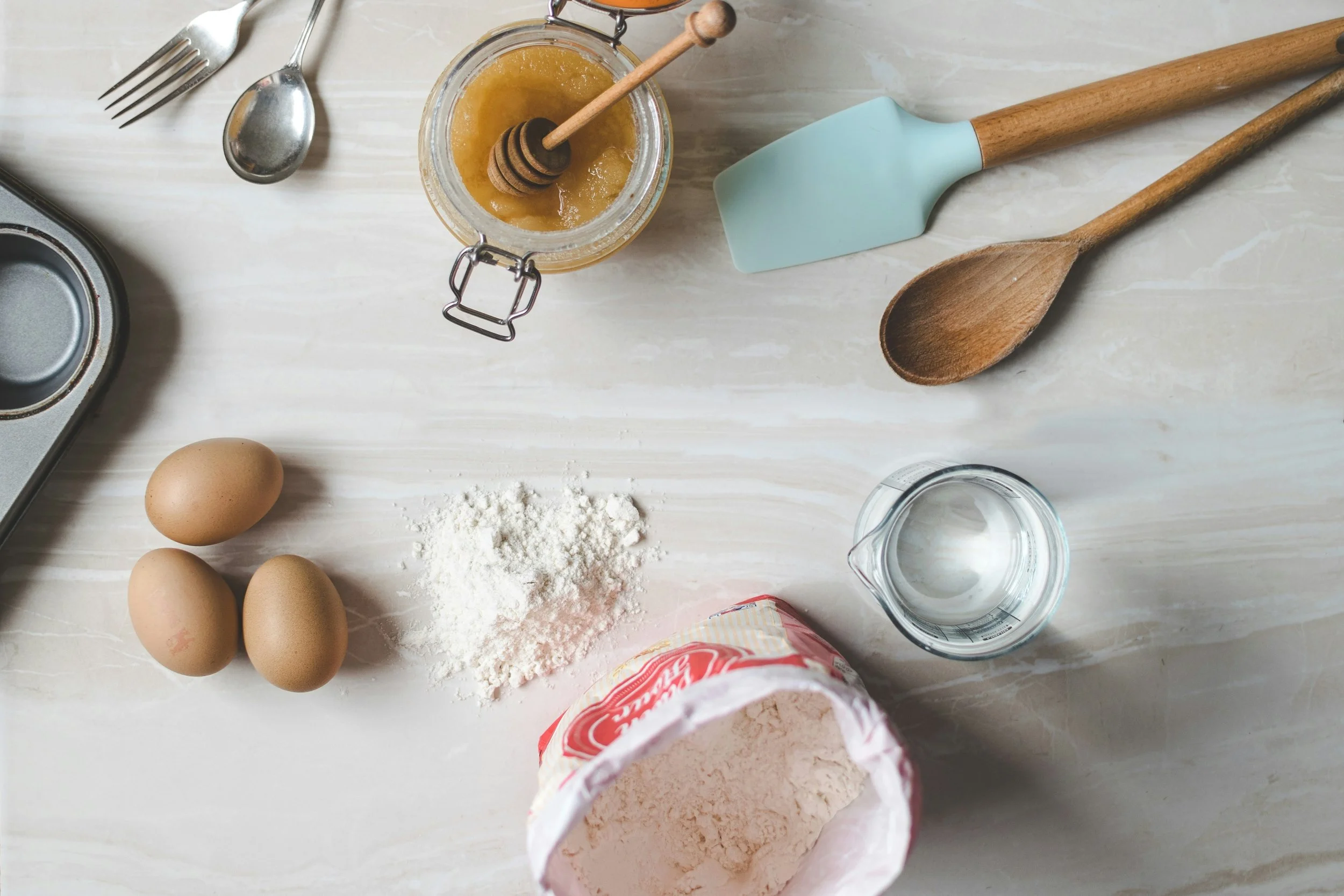 Kitchen countertop with eggs, flour, honey, water, and baking tools including a spatula, wooden spoons, and a honey dipper.