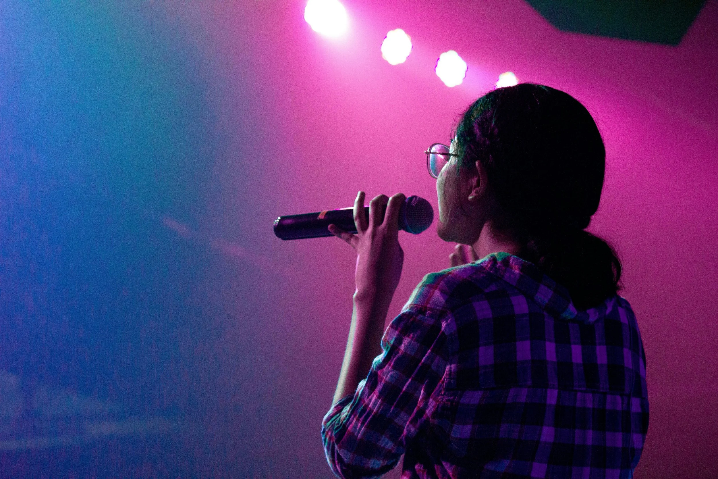 A woman wearing glasses and a plaid shirt singing into a microphone on a stage with pink and purple lighting.