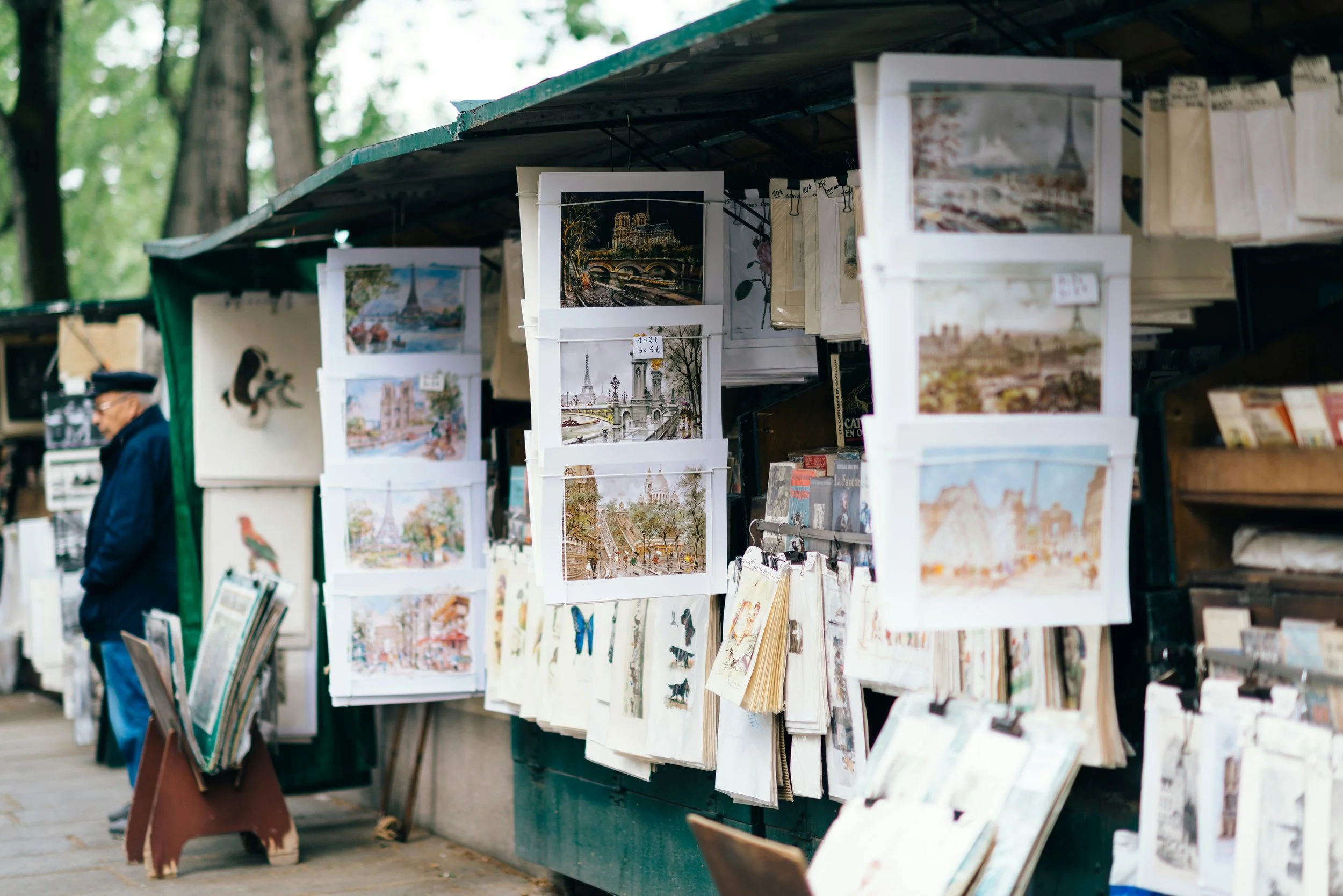 Outdoor street art and postcard stall with various travel postcards hanging on display, and a man browsing in the background.