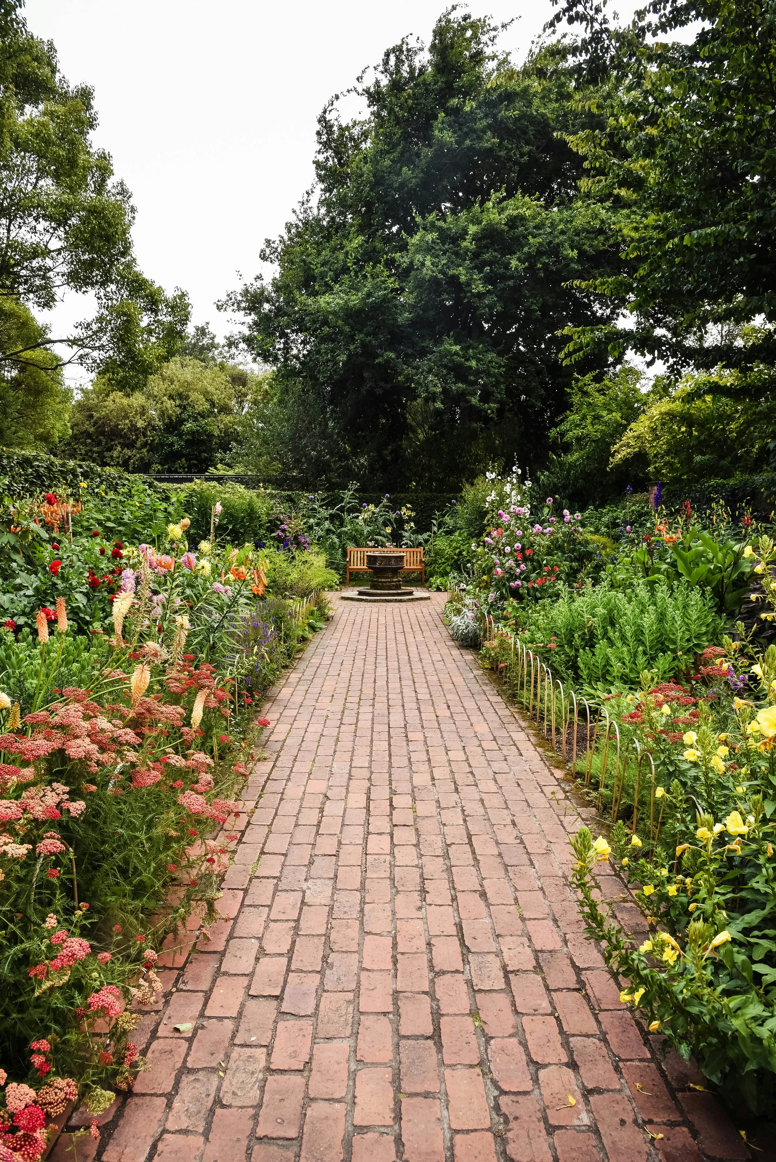 A garden pathway made of red bricks lined with colorful flowers on both sides, leading to a wooden bench and fountain in the distance, with lush green trees overhead. Curated Event Listings in Columbia, Maryland