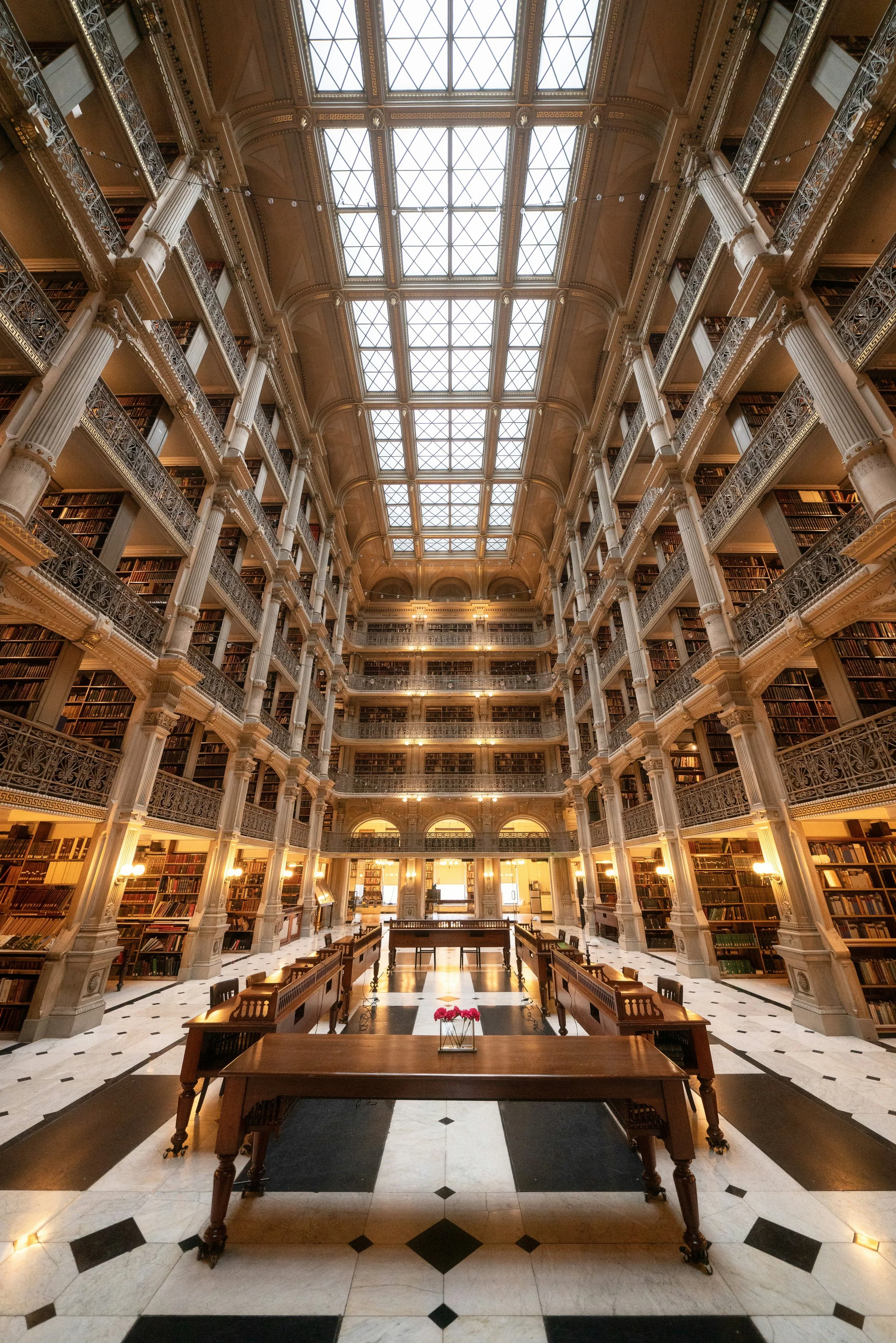 Interior view of a grand, historic library with multiple floors of bookshelves, ornate railings, chandeliers, and a glass-paned ceiling letting in natural light.