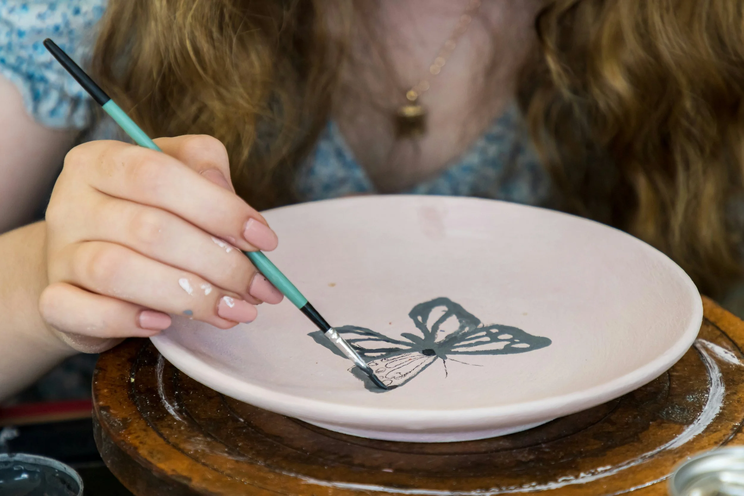 A woman painting a butterfly on a pink ceramic plate with a small brush.
