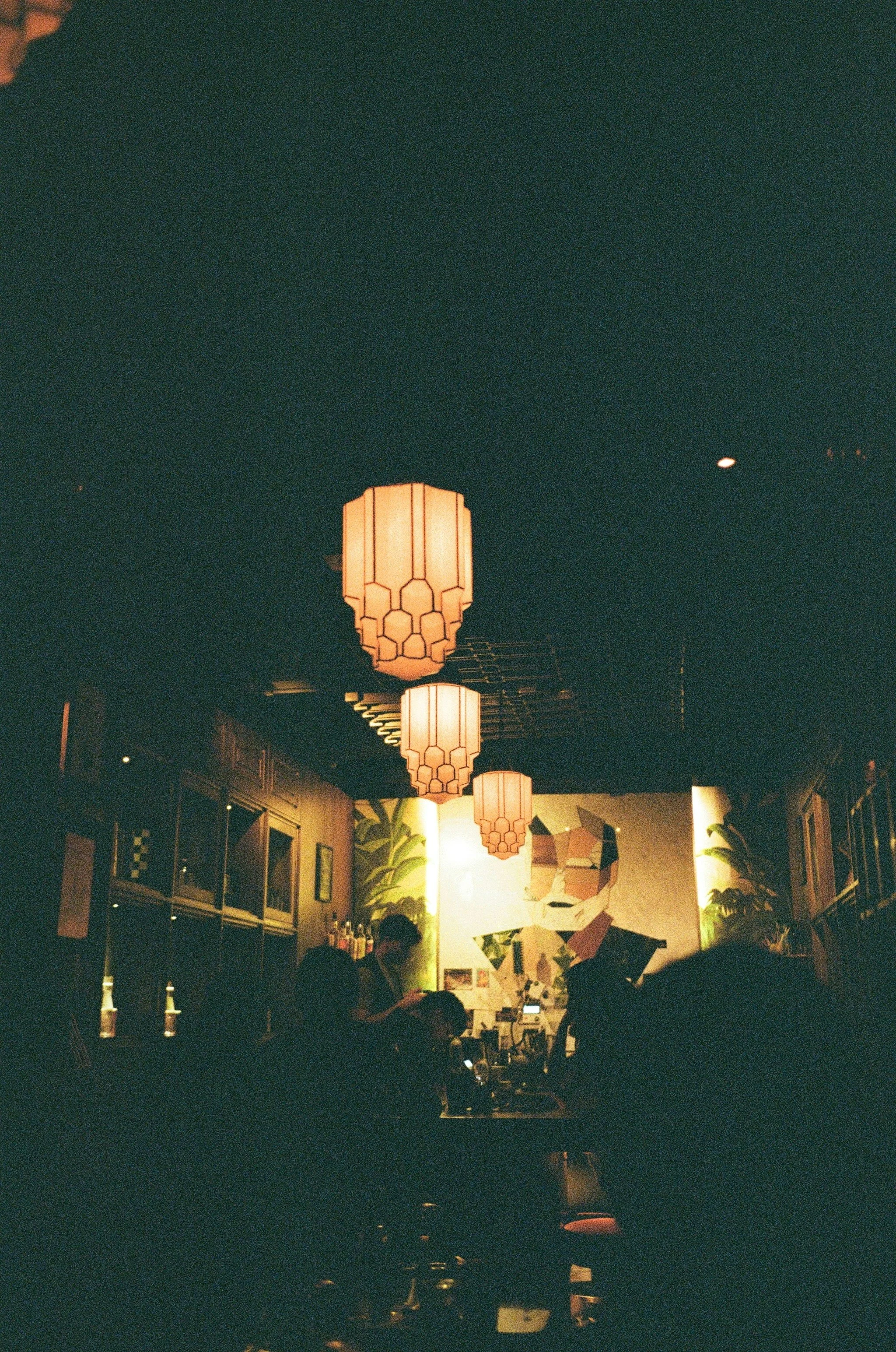 Dimly lit restaurant interior with hanging lanterns, people seated at the bar, and colorful abstract art on the wall.