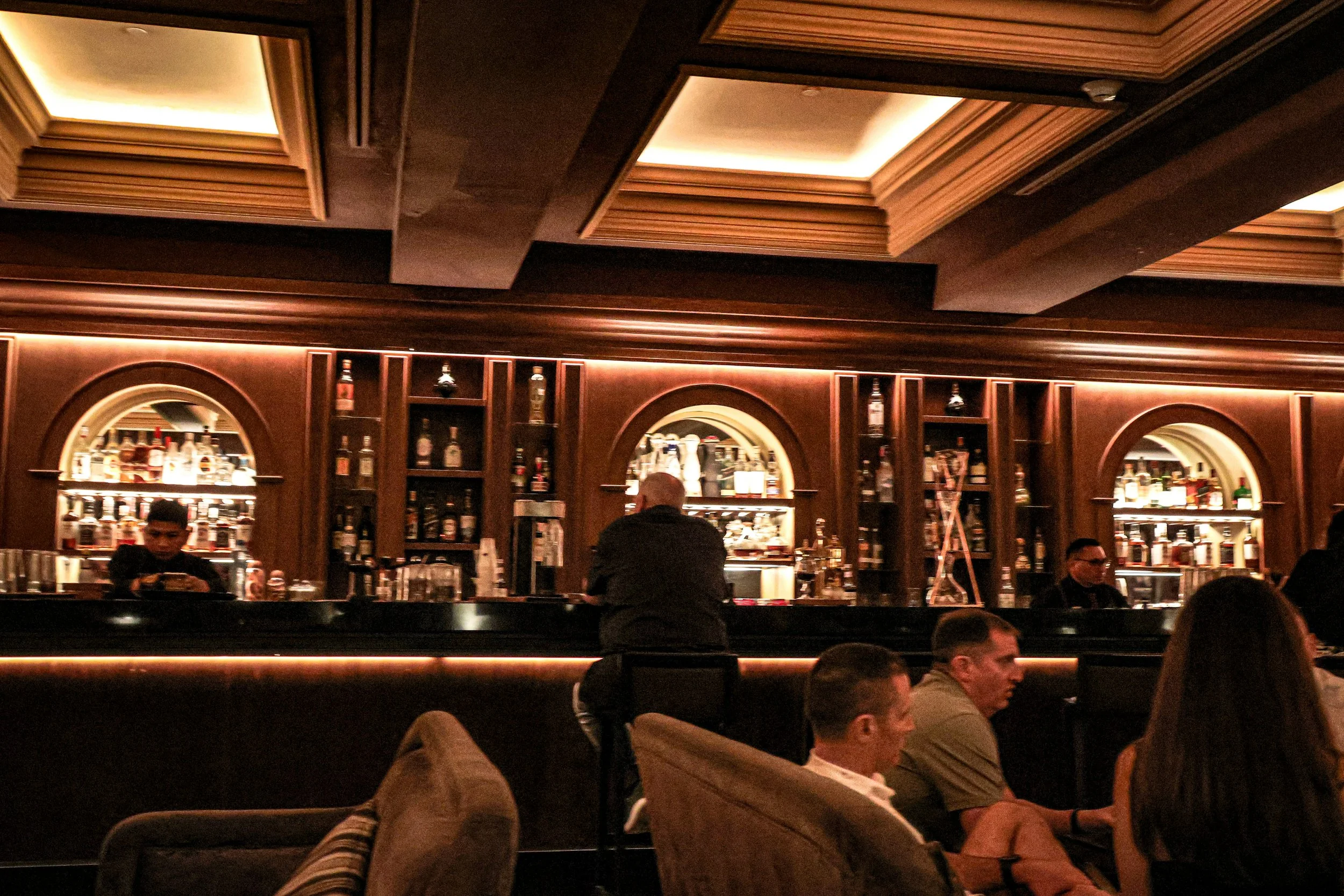 Interior view of a bar lounge with wooden shelves filled with liquor bottles, a marble countertop, and patrons sitting on couches and chairs, with some socializing and one person leaning against the bar.