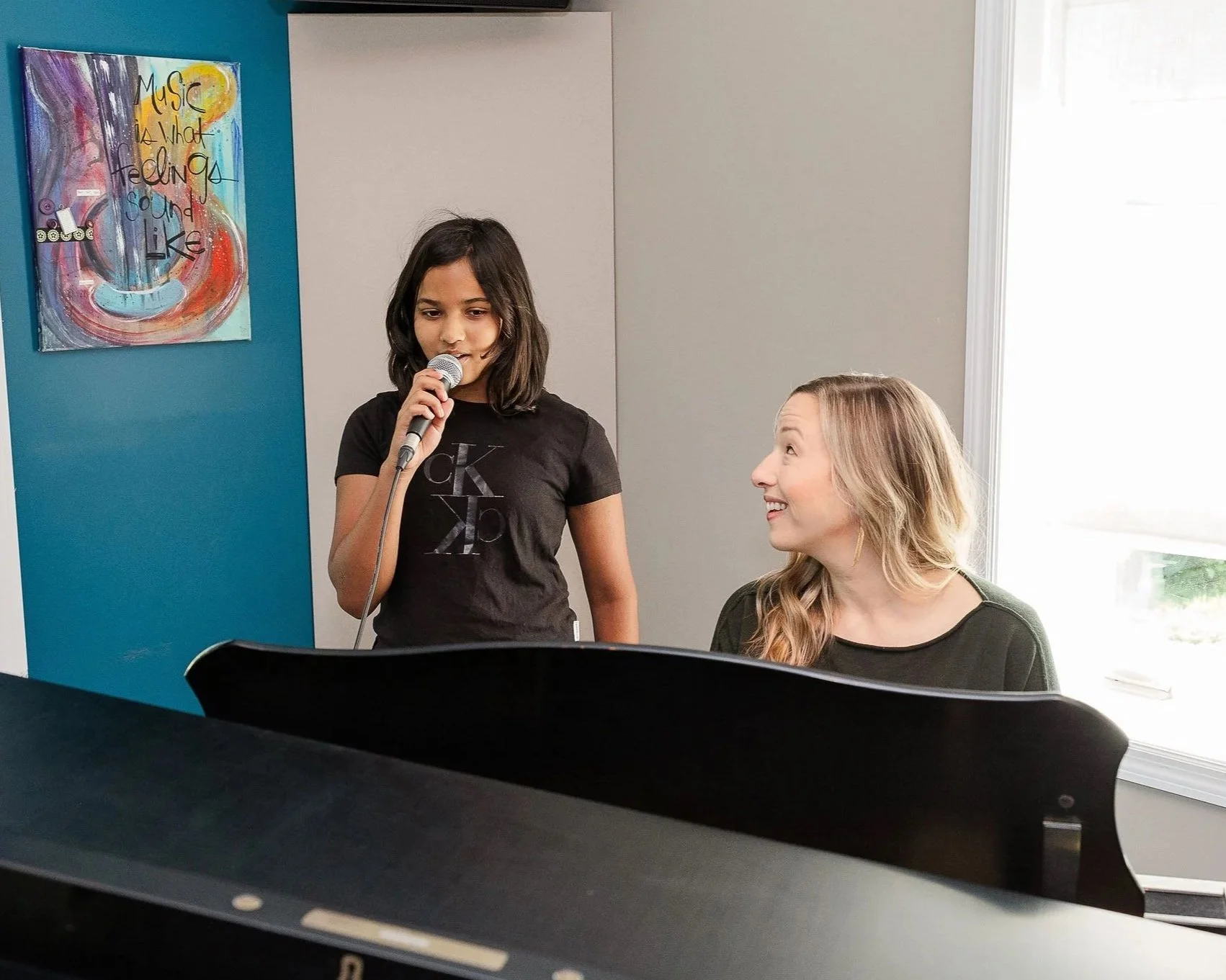 A young girl singing into a microphone while a woman, likely her music teacher or instructor, listens and smiles. They are in a music room with a colorful abstract painting on the wall that says, 'Music is what feeling sound like.'