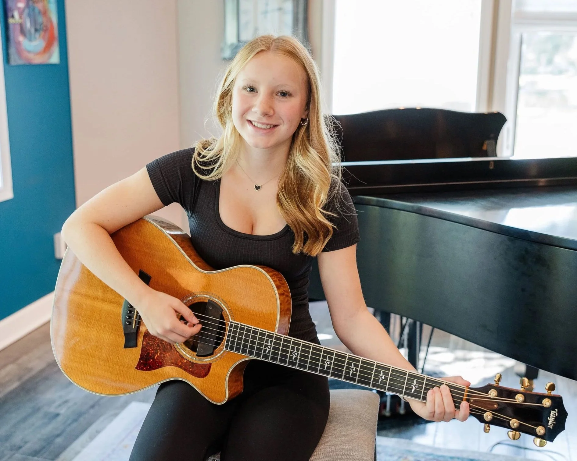 A young woman with long, wavy blonde hair sitting on a beige bench, playing an acoustic guitar, smiling, in a well-lit room with windows and a black piano in the background.