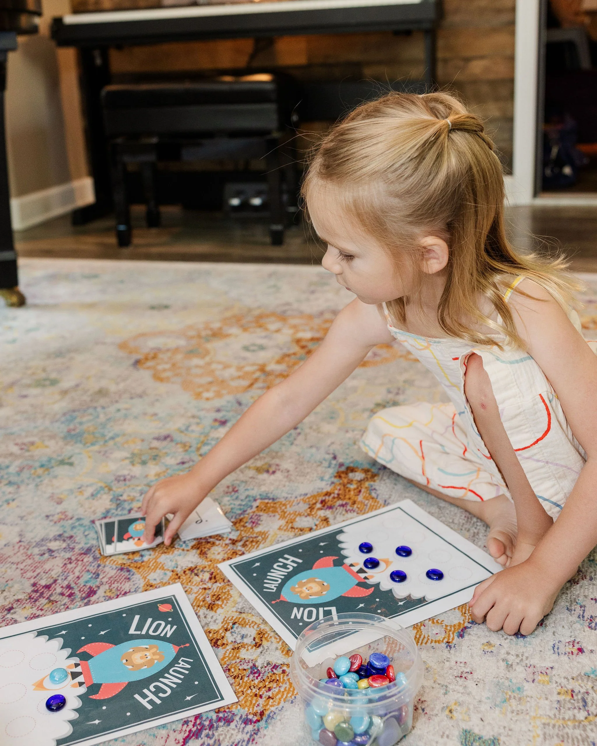 A young girl with blonde hair playing a music piano game called 'Launch Lion' on a colorful rug. She is reaching for game cards while seated on the music studio rug in the music lesson studio in Lake Elmo.