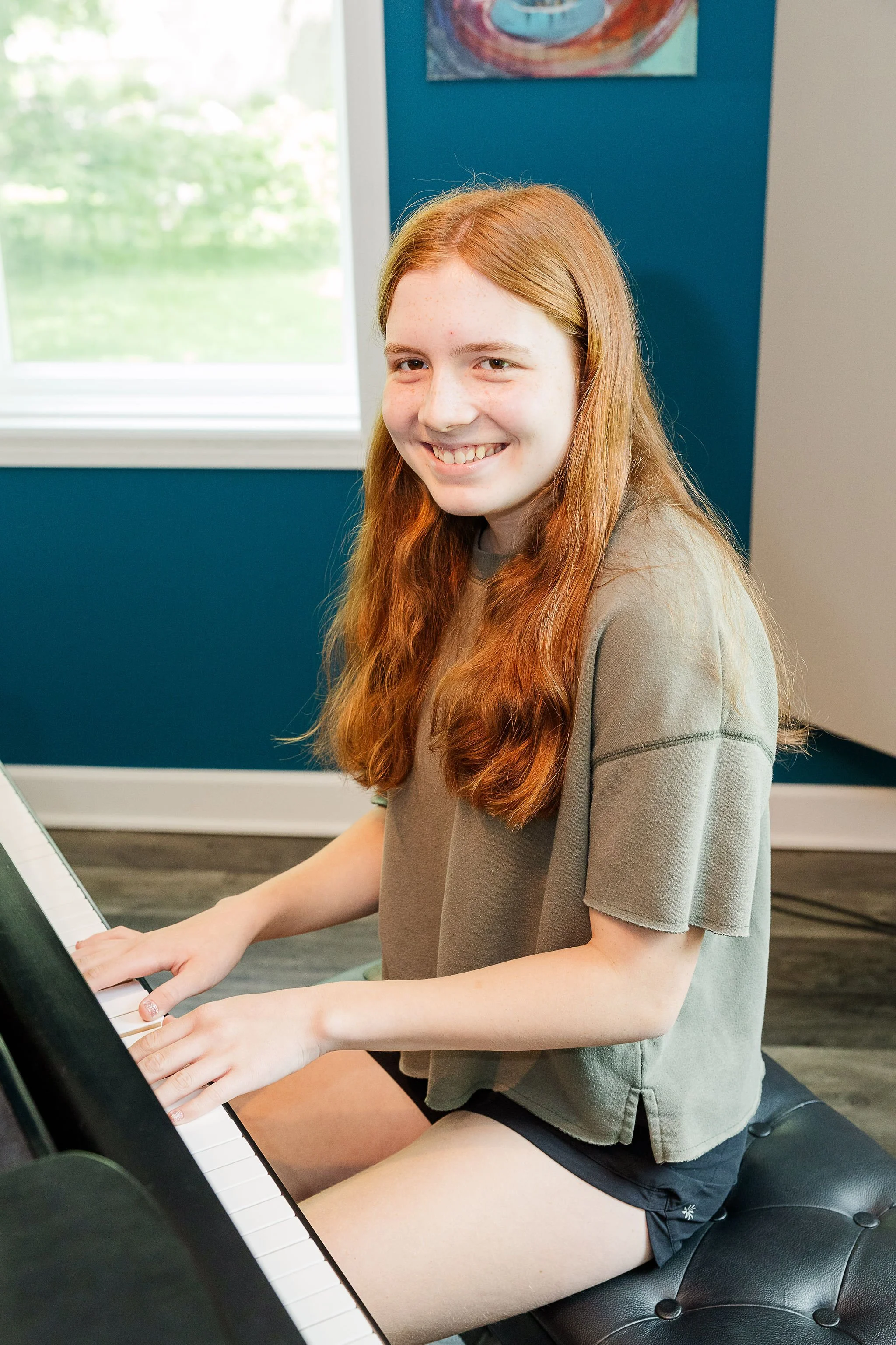 A teenage girl with long red hair and a gray T-shirt playing the piano indoors, smiling at the camera.