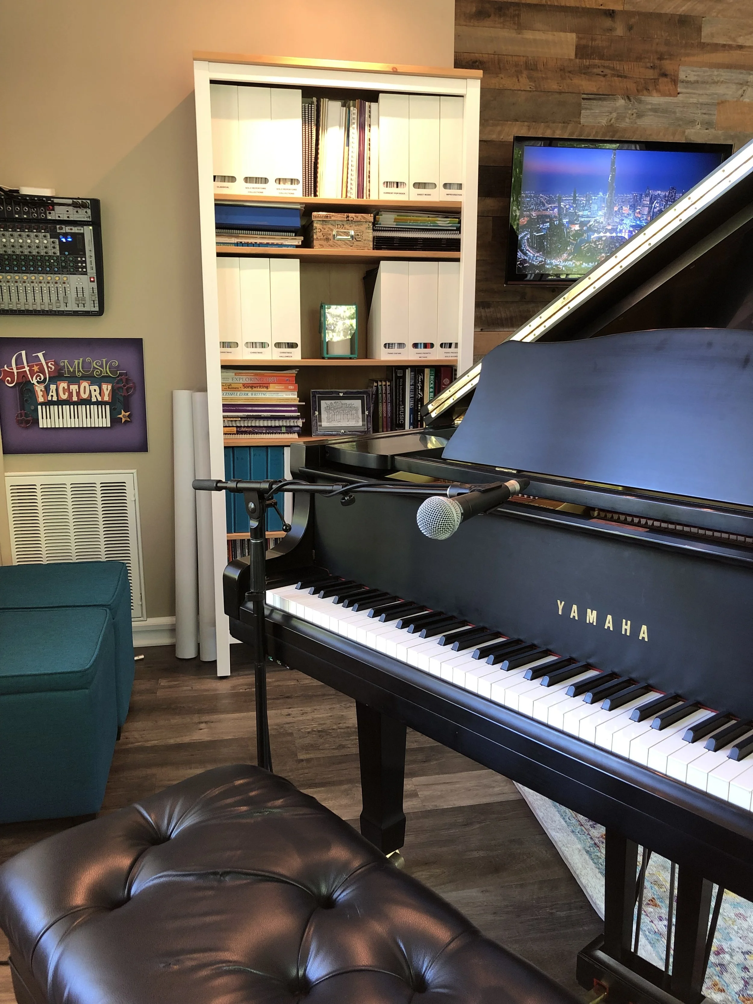 A black Yamaha grand piano with a microphone on a stand in front of it, in a room with a bookshelf, a teal couch, a TV displaying a cityscape at night, and various other musical equipment and decor.