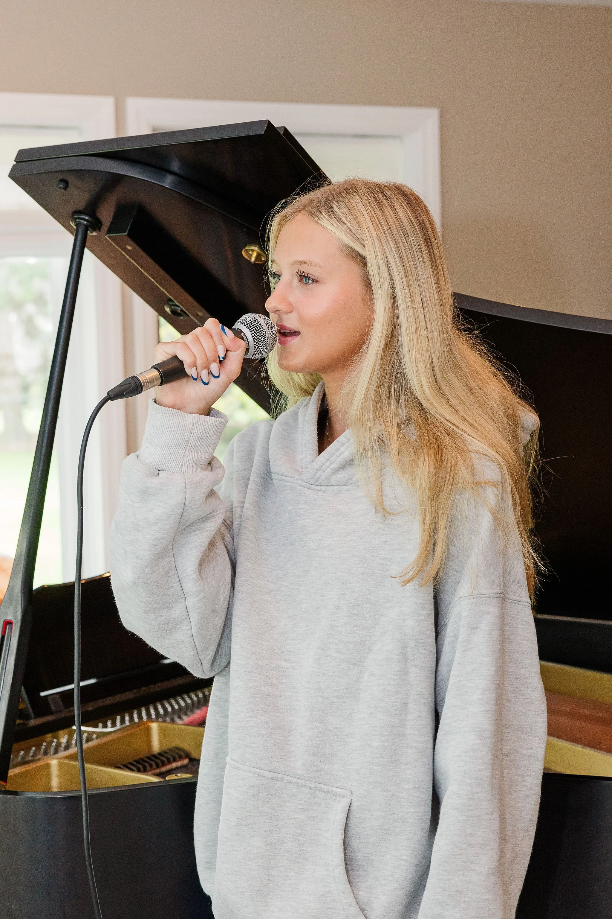 A young woman with blonde hair singing into a microphone next to a grand piano.