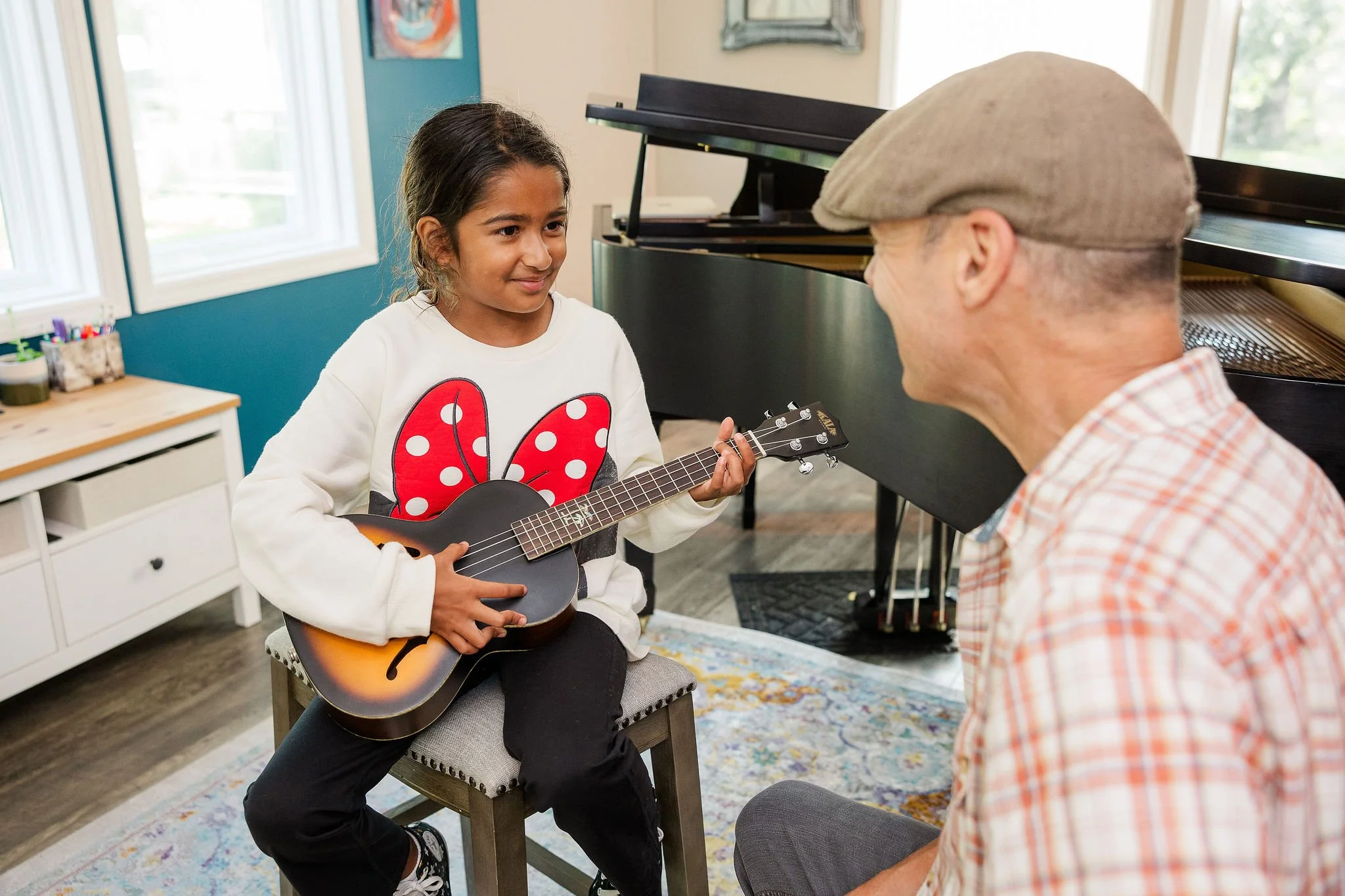 A young girl sitting on a chair, holding a ukulele, and looking at a man who is smiling at her. The girl is wearing a white shirt with a red polka dot bow design, and the man is wearing a plaid shirt and a brown cap. They are in a room with a piano a