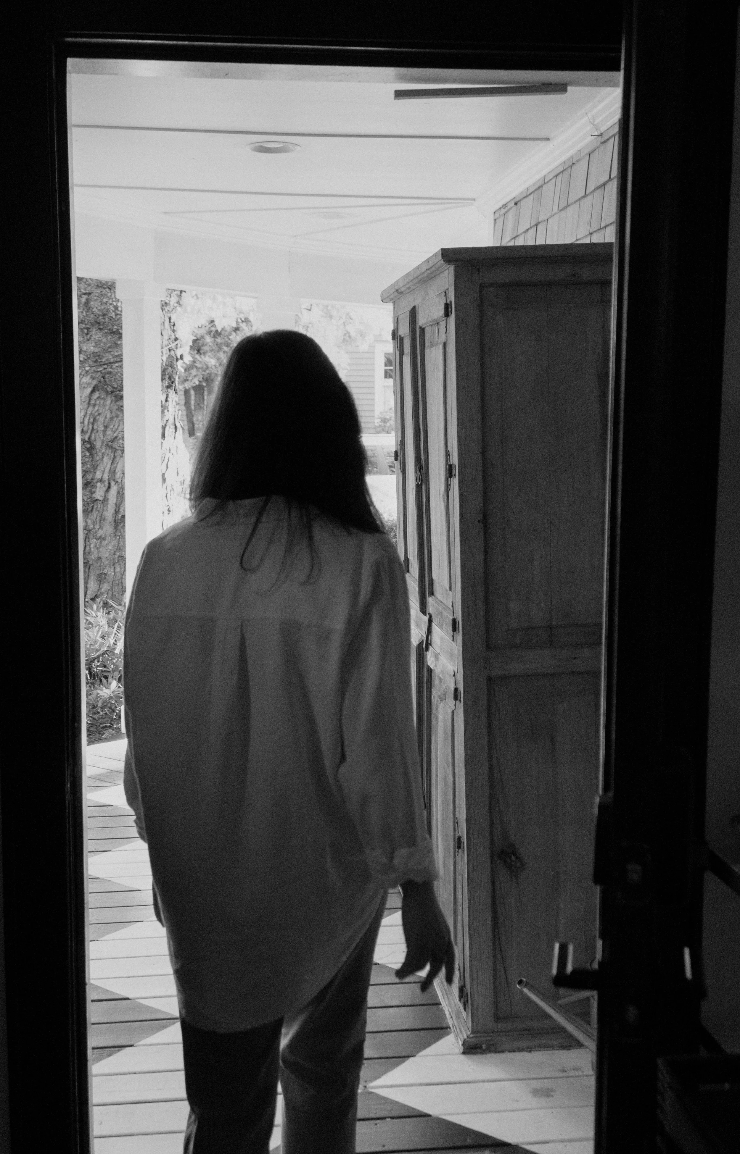 A woman with dark hair wearing a light-colored shirt walking out of a house onto a porch, with greenery and trees visible outside.