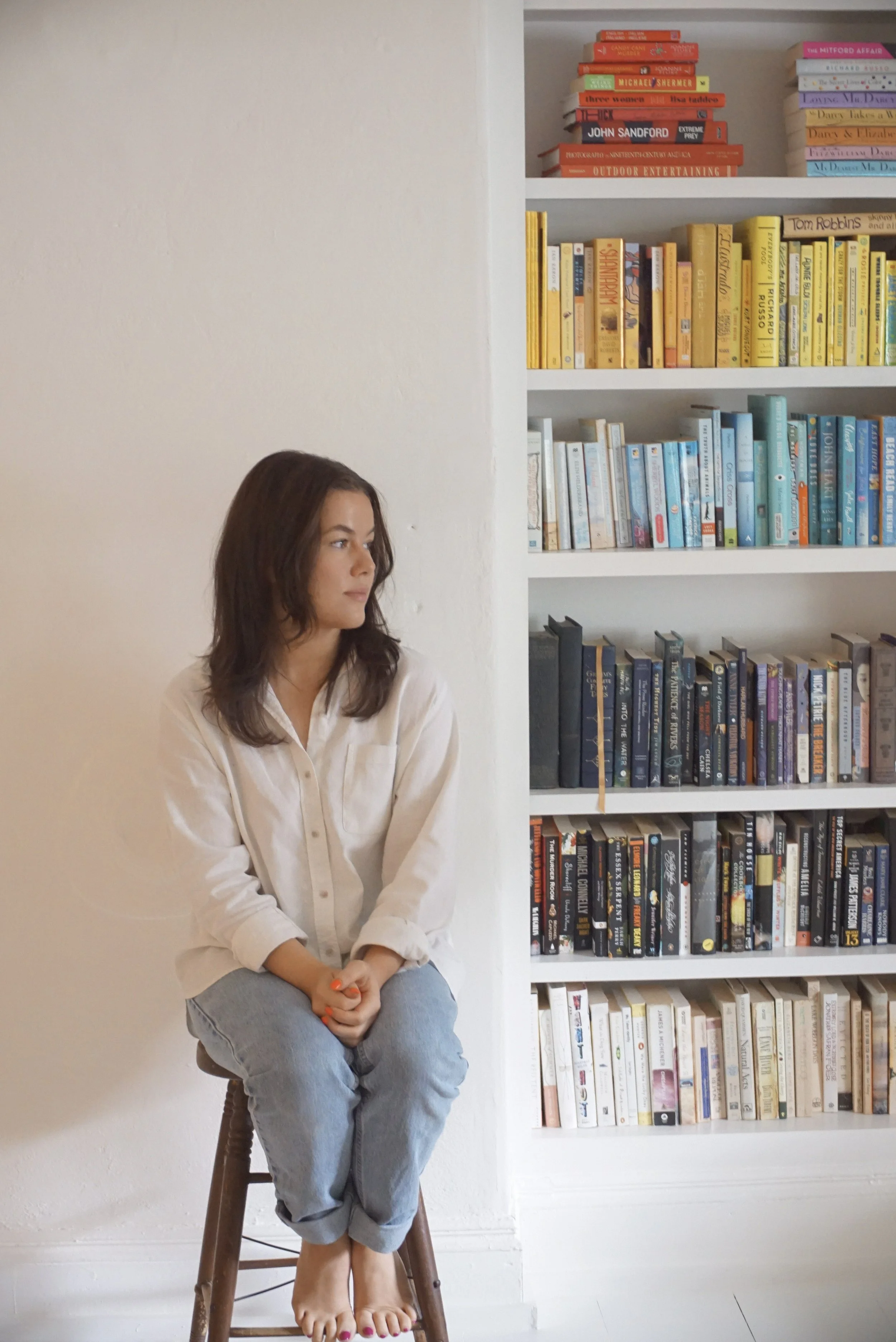 A woman sits on a wooden chair with her hands clasped on her lap, positioned in front of a white wall to the left and a white bookshelf filled with colorful books to the right.