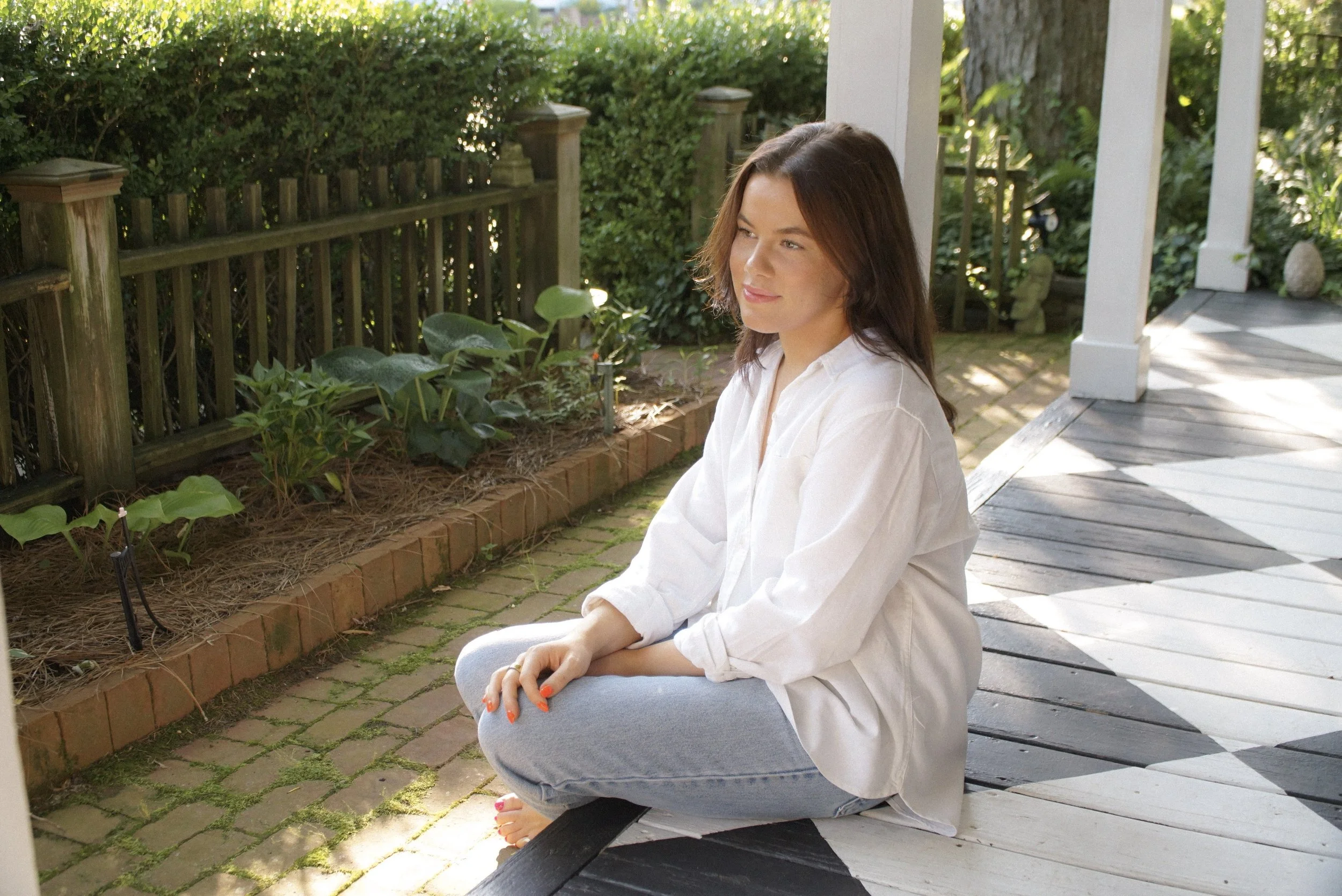 A woman with brown hair wearing a white shirt and light blue jeans, sitting cross-legged on a porch near a garden with green plants and a wooden fence, enjoying sunny weather.