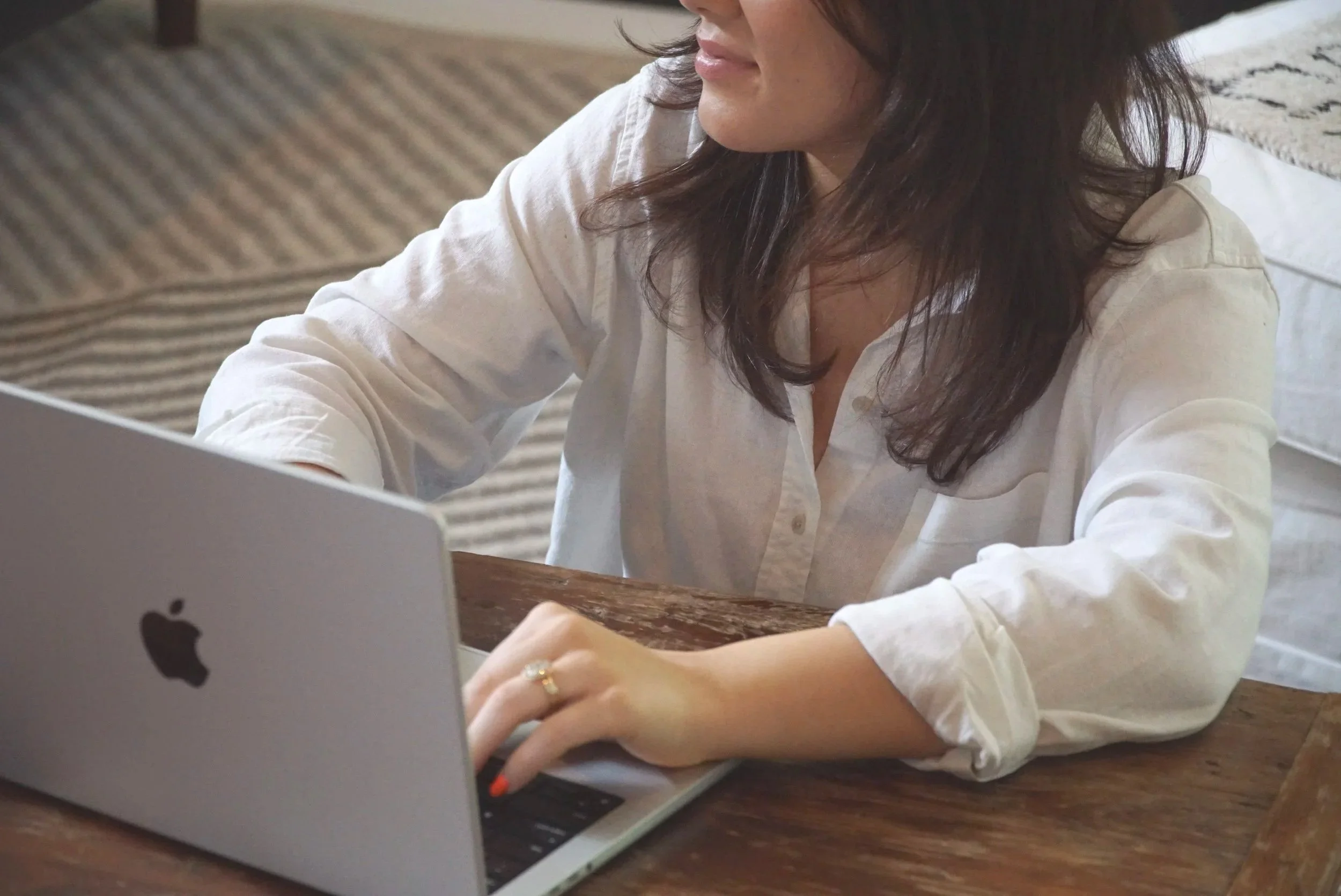 Woman with dark hair wearing a white shirt working on a silver MacBook at a wooden table.