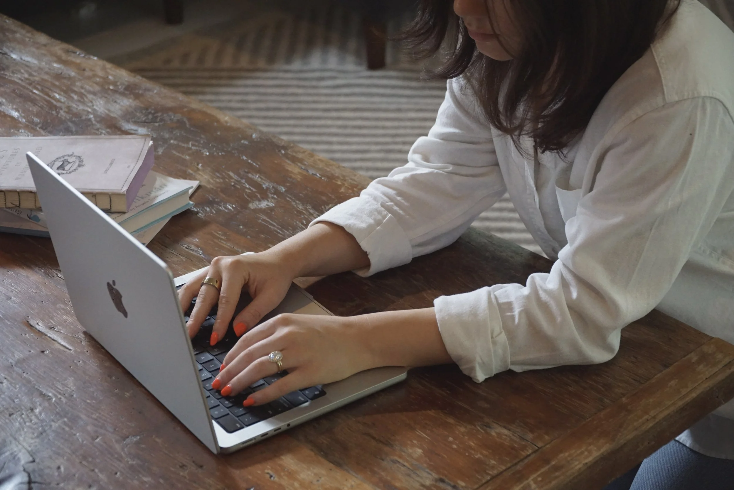 A woman typing on a silver MacBook on a wooden table, with books nearby.