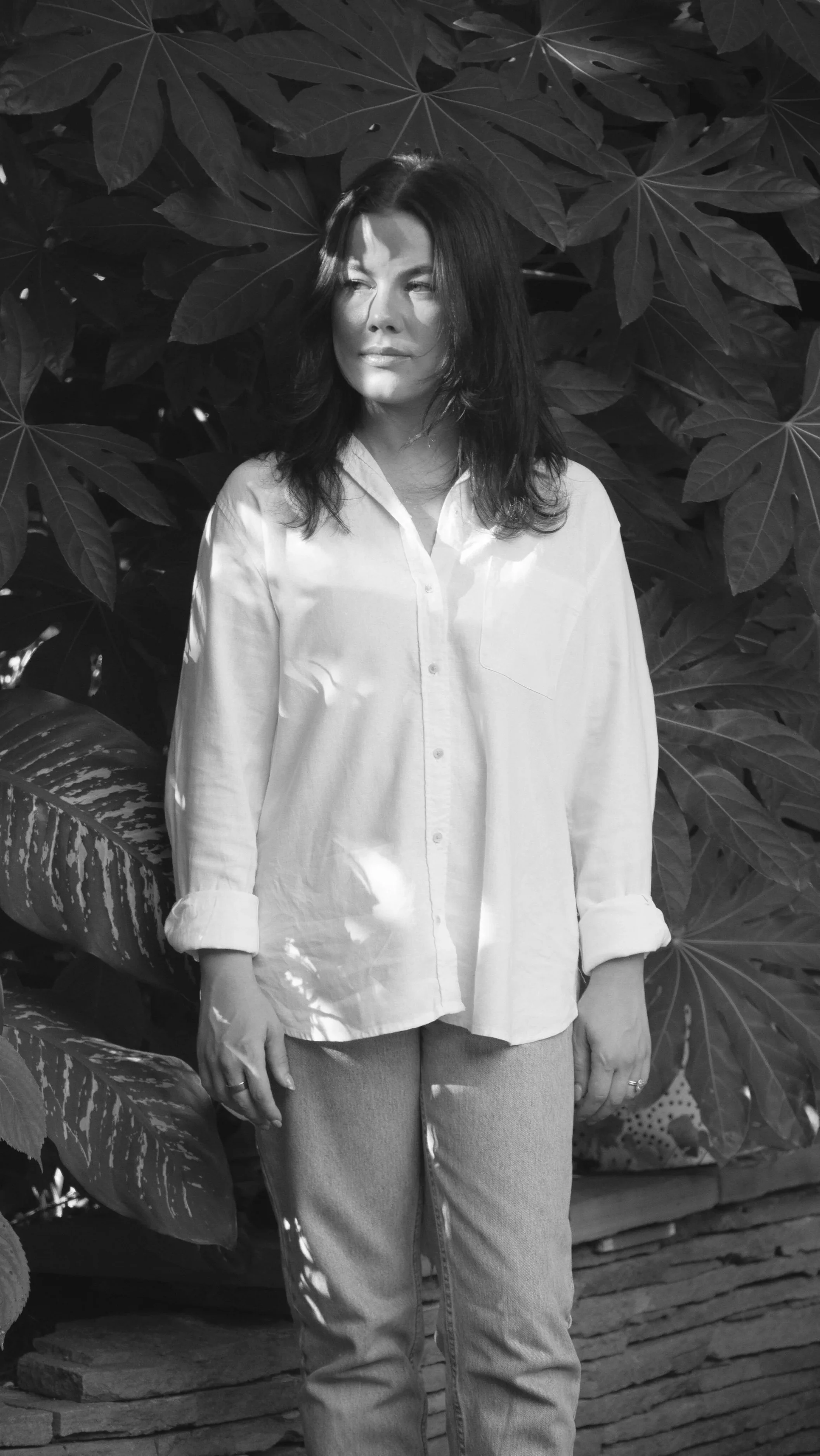 A woman with dark hair in a white button-up shirt and jeans standing outdoors amidst large leafy plants.