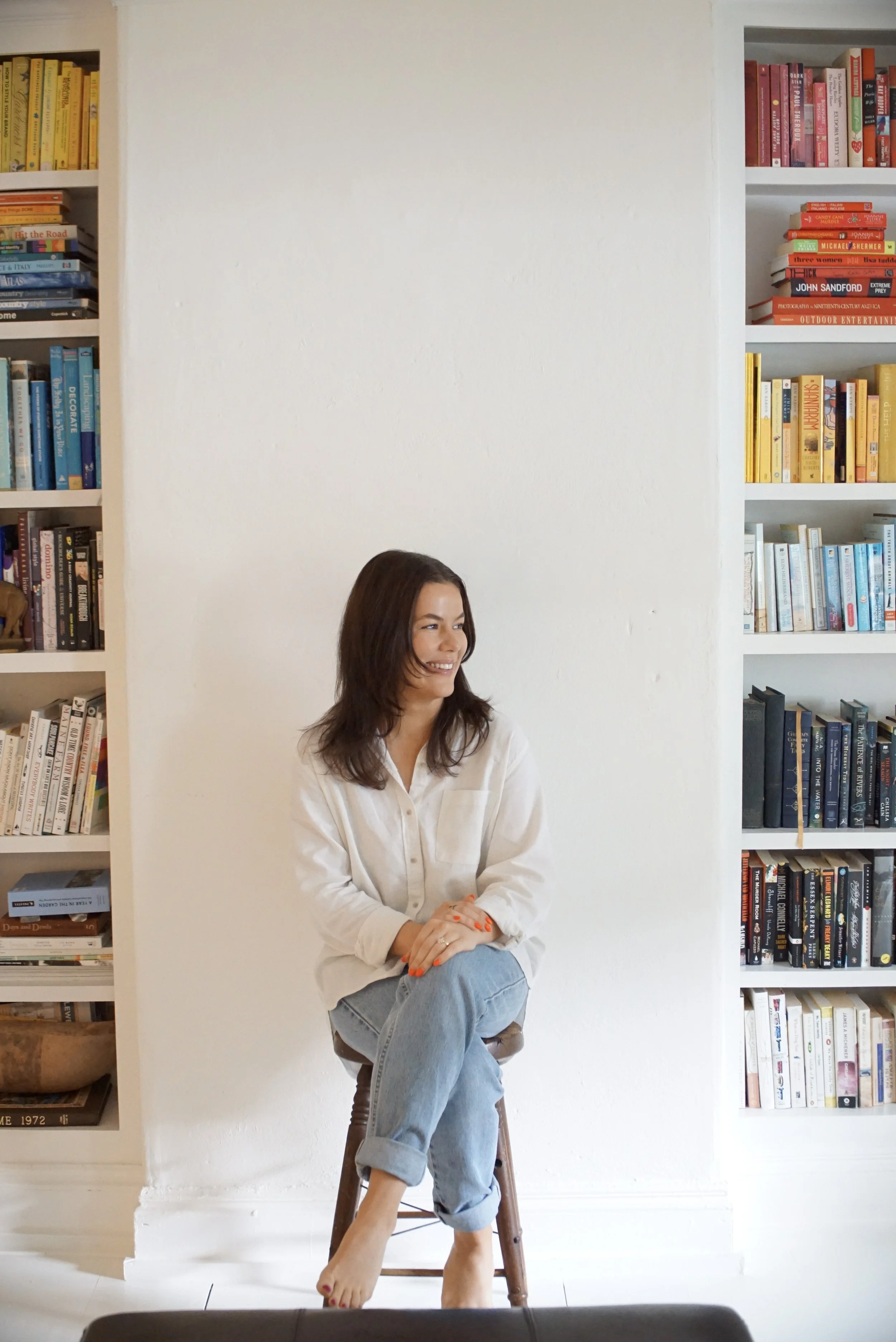 A woman with shoulder-length brown hair, wearing a white blouse and light blue jeans, sitting on a wooden stool in front of a white wall surrounded by built-in bookshelves filled with colorful books.