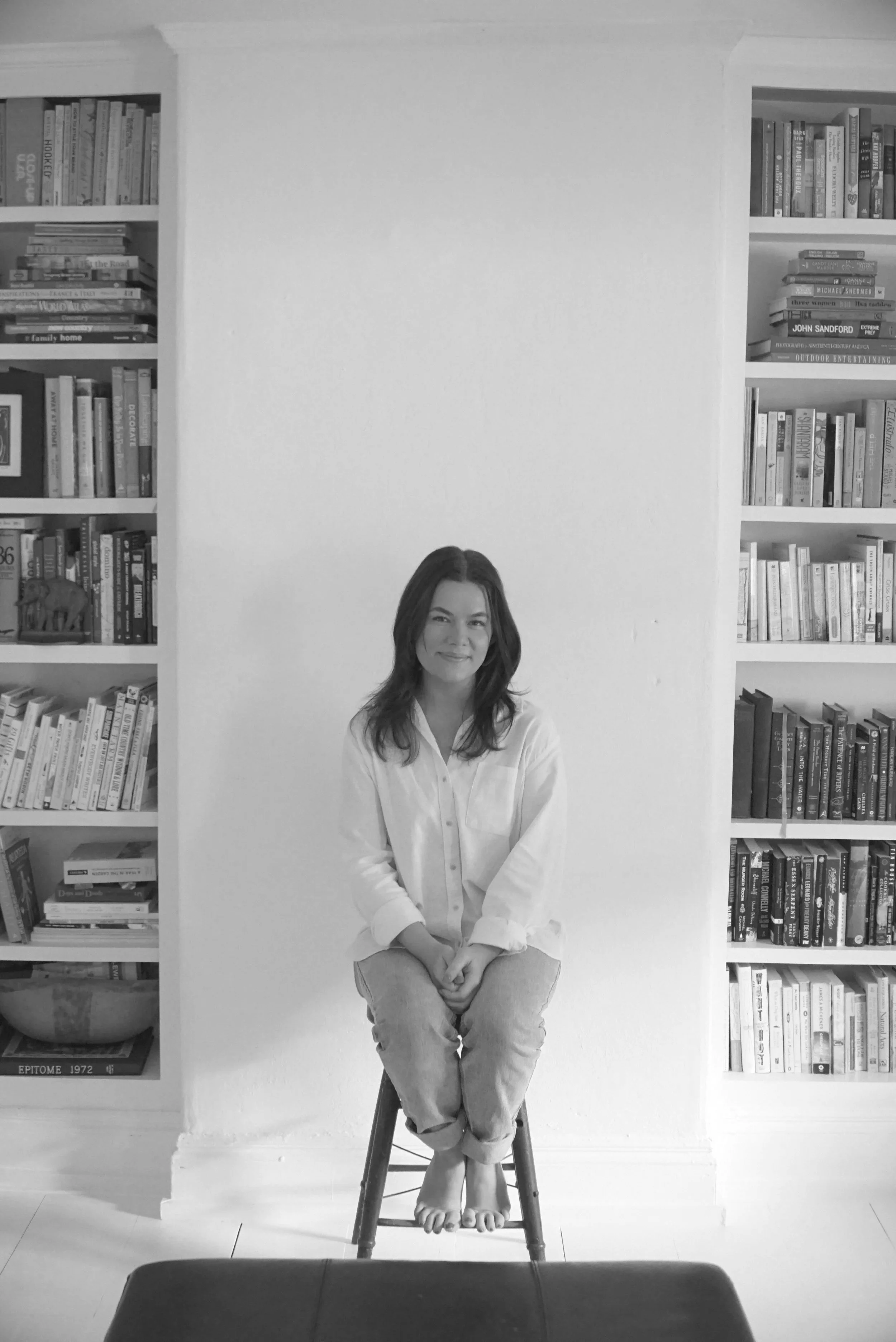 A woman sitting on a stool with her feet on the rung, in front of a white wall and flanked by built-in bookshelves filled with books.