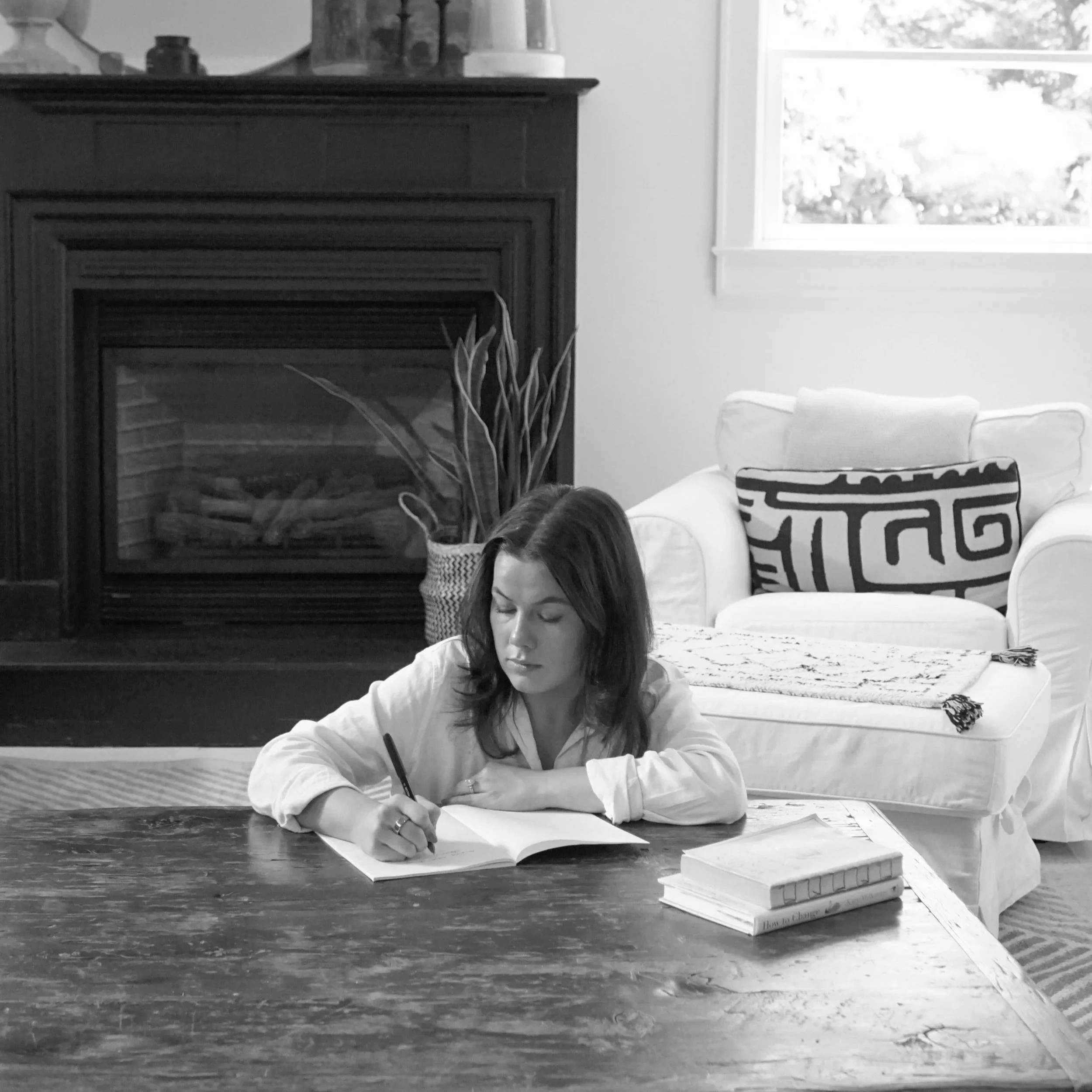 A woman with shoulder-length dark hair sits at a wooden table, writing in a notebook with a pen. There are a few books stacked on the table to her right. Behind her is a white armchair with a patterned pillow and a throw blanket. A fireplace with a tall plant in a woven basket is visible in the background, along with a window letting in natural light.