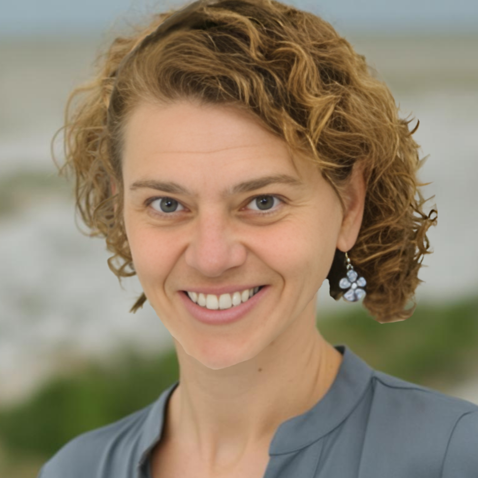 Close-up of a woman with short, curly red hair and blue eyes, smiling outdoors with blurred greenery and sky in the background.