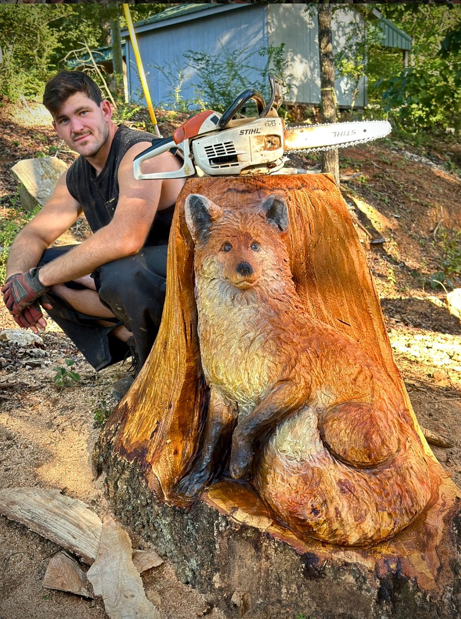 A young man kneeling next to a wood sculpture of a dog carved into a tree trunk, with a chainsaw resting on top of the sculpture. The scene is outdoors with trees and a small building in the background.