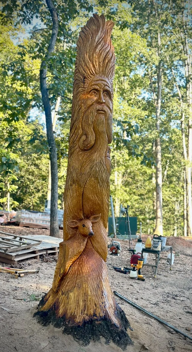 A carved wooden totem pole featuring a man's face with long hair and a beard, a bear, and a smaller animal, set in a wooded outdoor area with tools and equipment nearby.