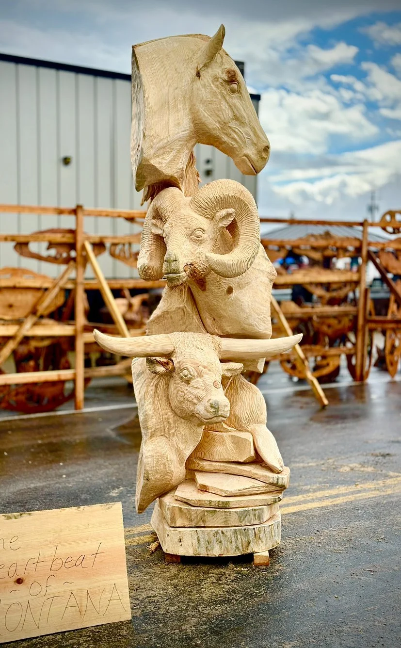 Wooden sculpture of a stack of four carved animals including a horse at the top, a ram, a buffalo, and a bison at the bottom, with a small wooden sign nearby.
