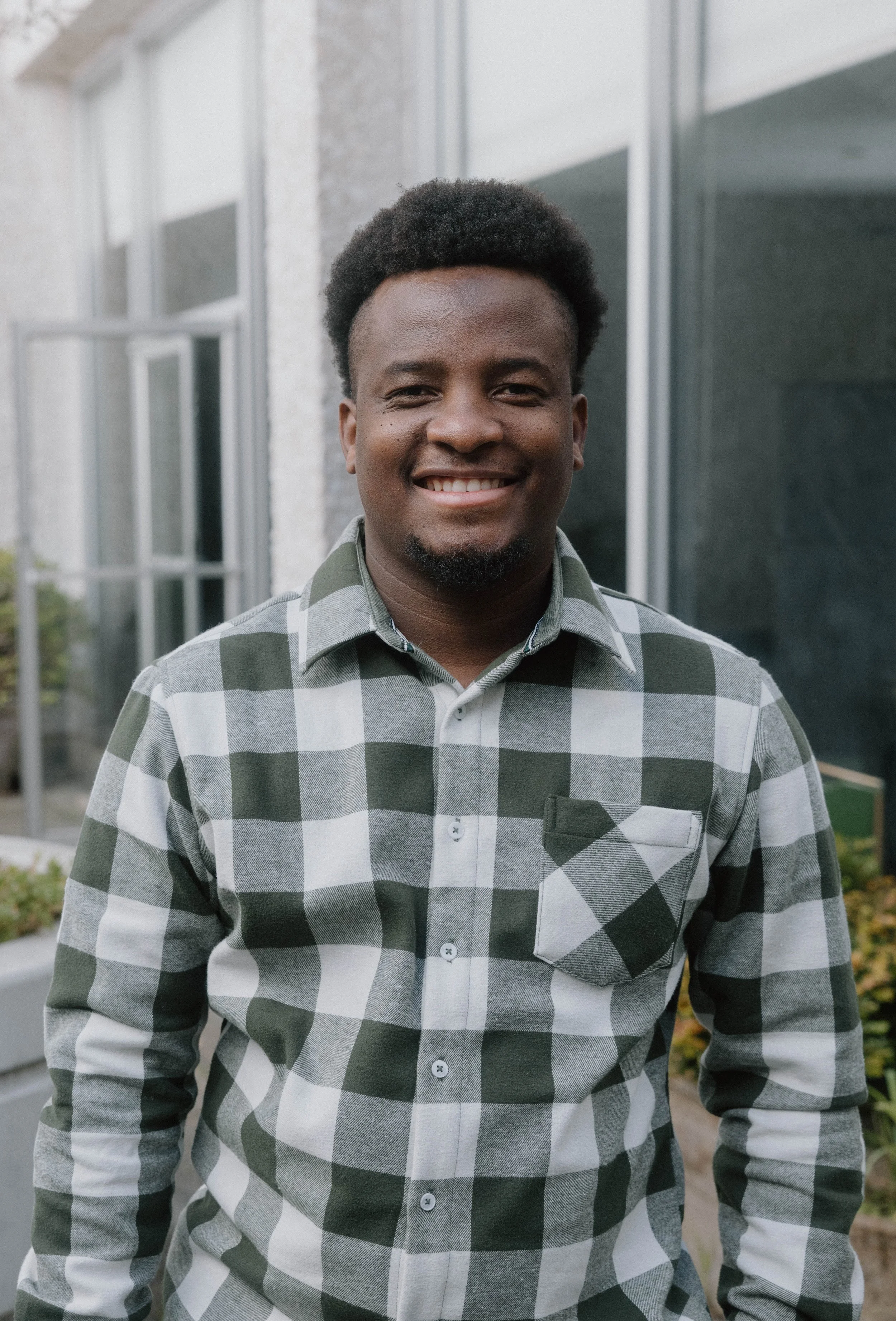 A man with dark skin, curly black hair, and a goatee, smiling and wearing a green and white checkered shirt, standing outside in front of a modern building with large glass windows.