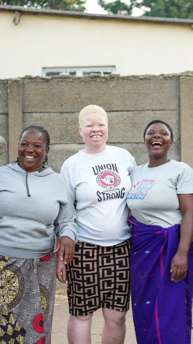 Three women standing outdoors, smiling and laughing together, with a concrete wall and a building in the background.
