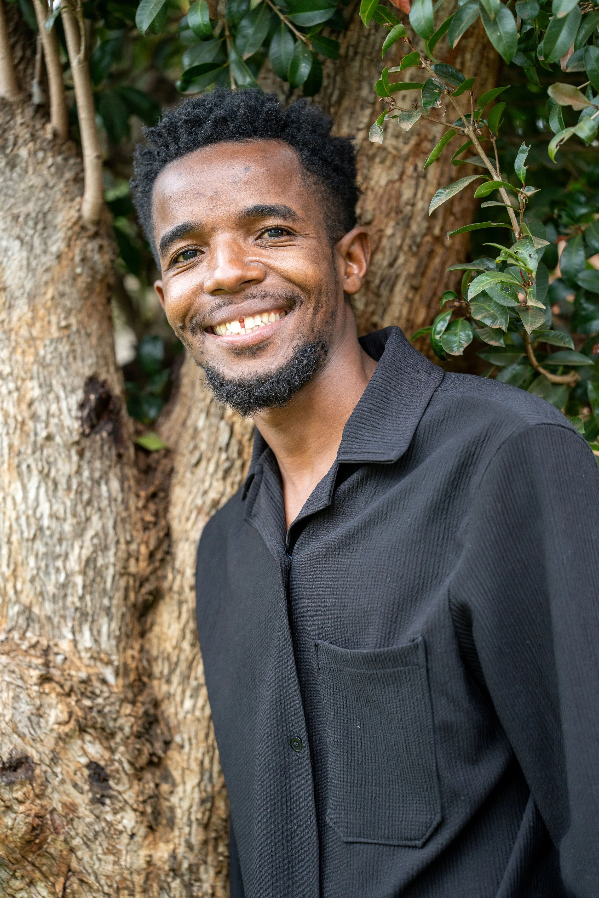 A man smiling, with short curly black hair and a beard, standing outdoors near a large tree with green leaves.