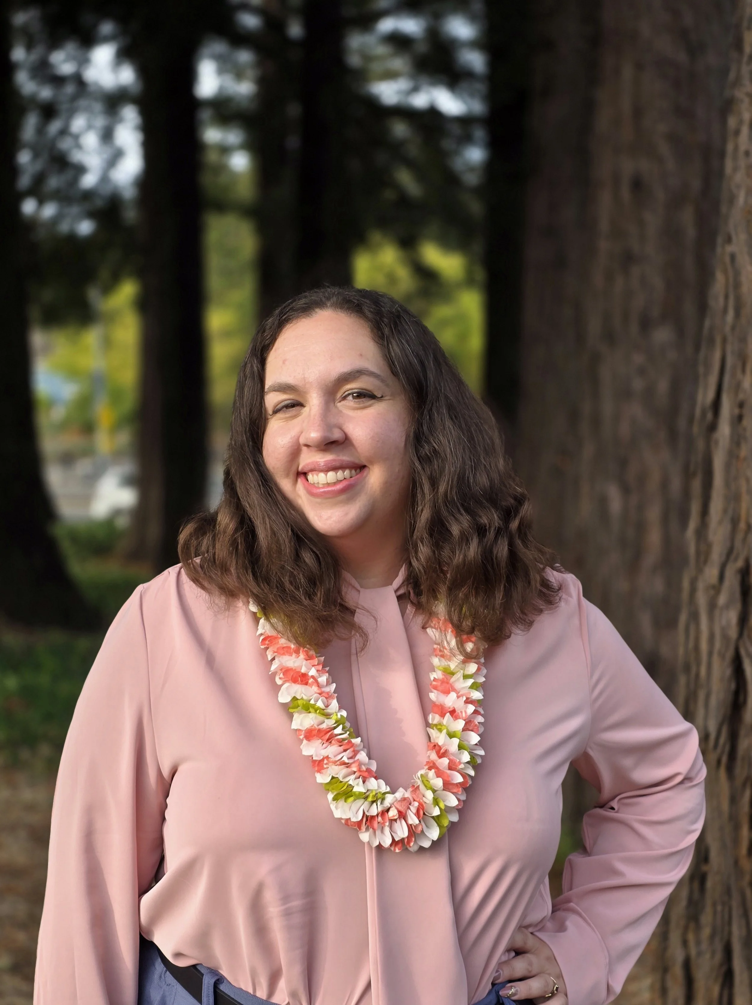 A woman with brown, curly hair smiling outdoors, wearing a pink dress and a colorful flower lei, standing in front of trees.