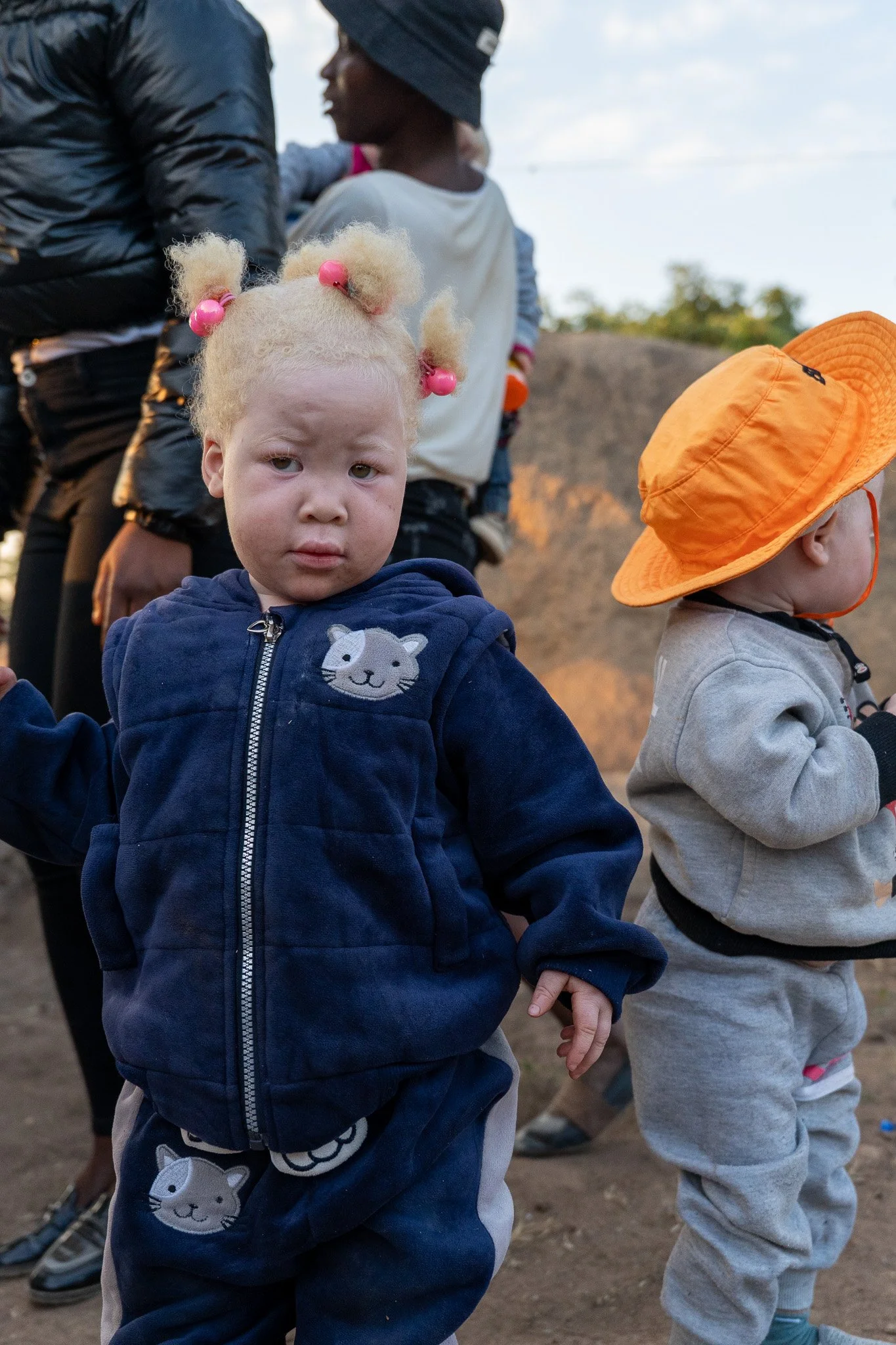 Two young children holding hands, one girl with blond hair styled in pigtails with pink beads, wearing a navy hoodie with cat face design, and a boy with short hair in a gray sweatshirt and orange bucket hat, outdoors during daytime.